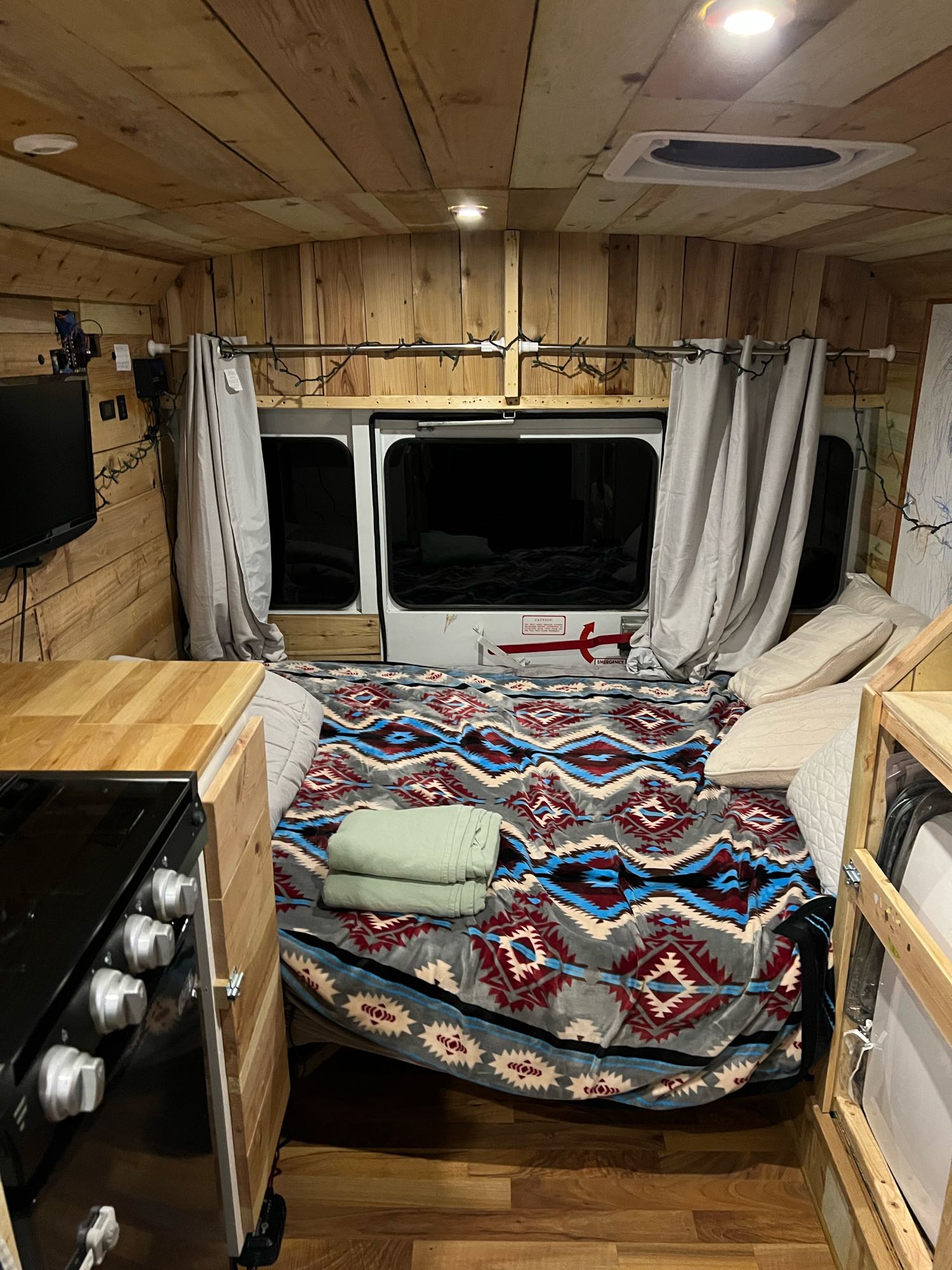 Cozy rv rental interior featuring wood-paneled walls, a comfy bed, kitchen area, and windows with curtains.. Chevy Express 3500 Mini Bus 2002