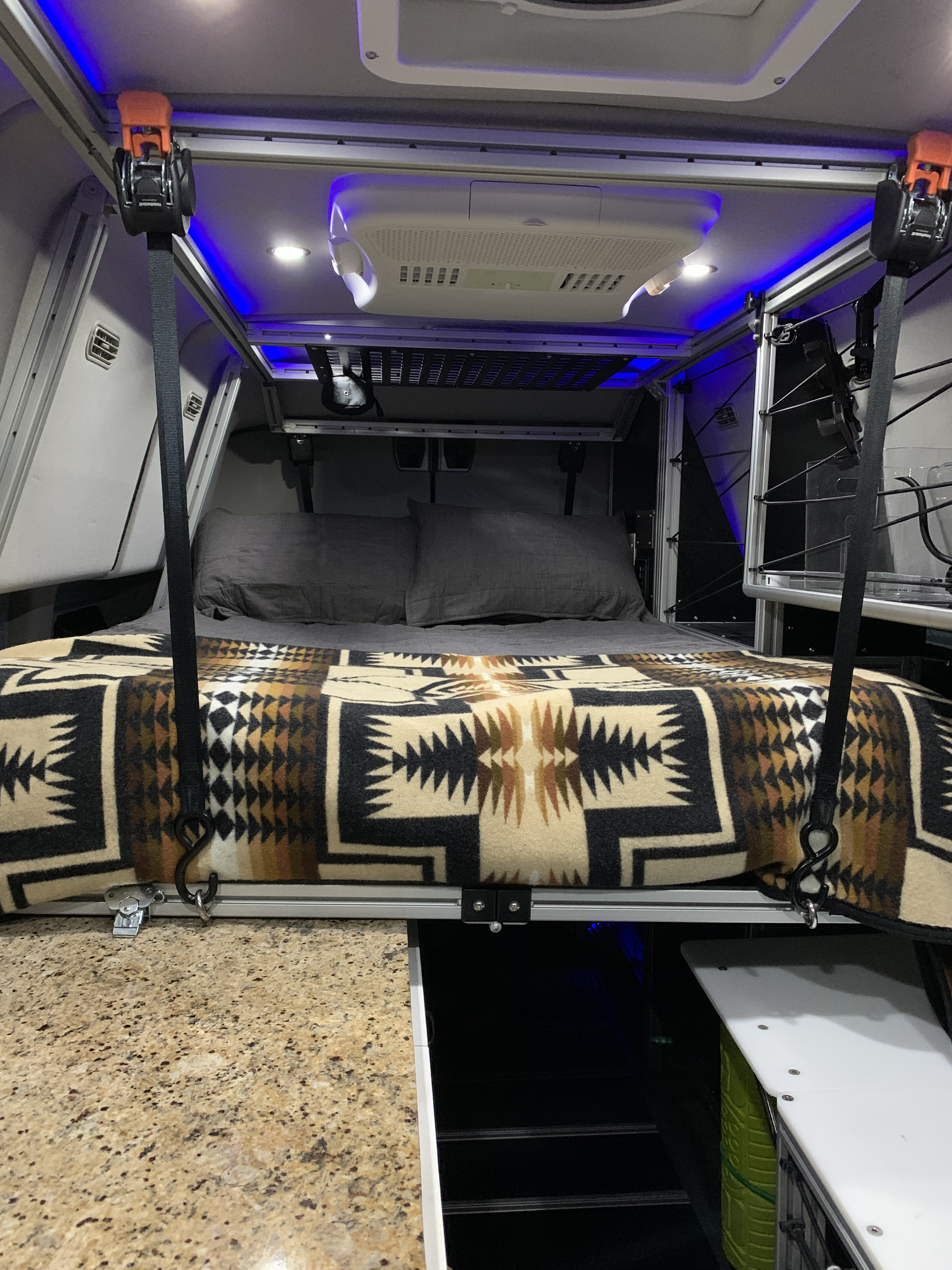 RV rental interior: A suspended bed with a patterned blanket, overhead AC, and blue lighting, maximizing space.. Ford Transit 2019