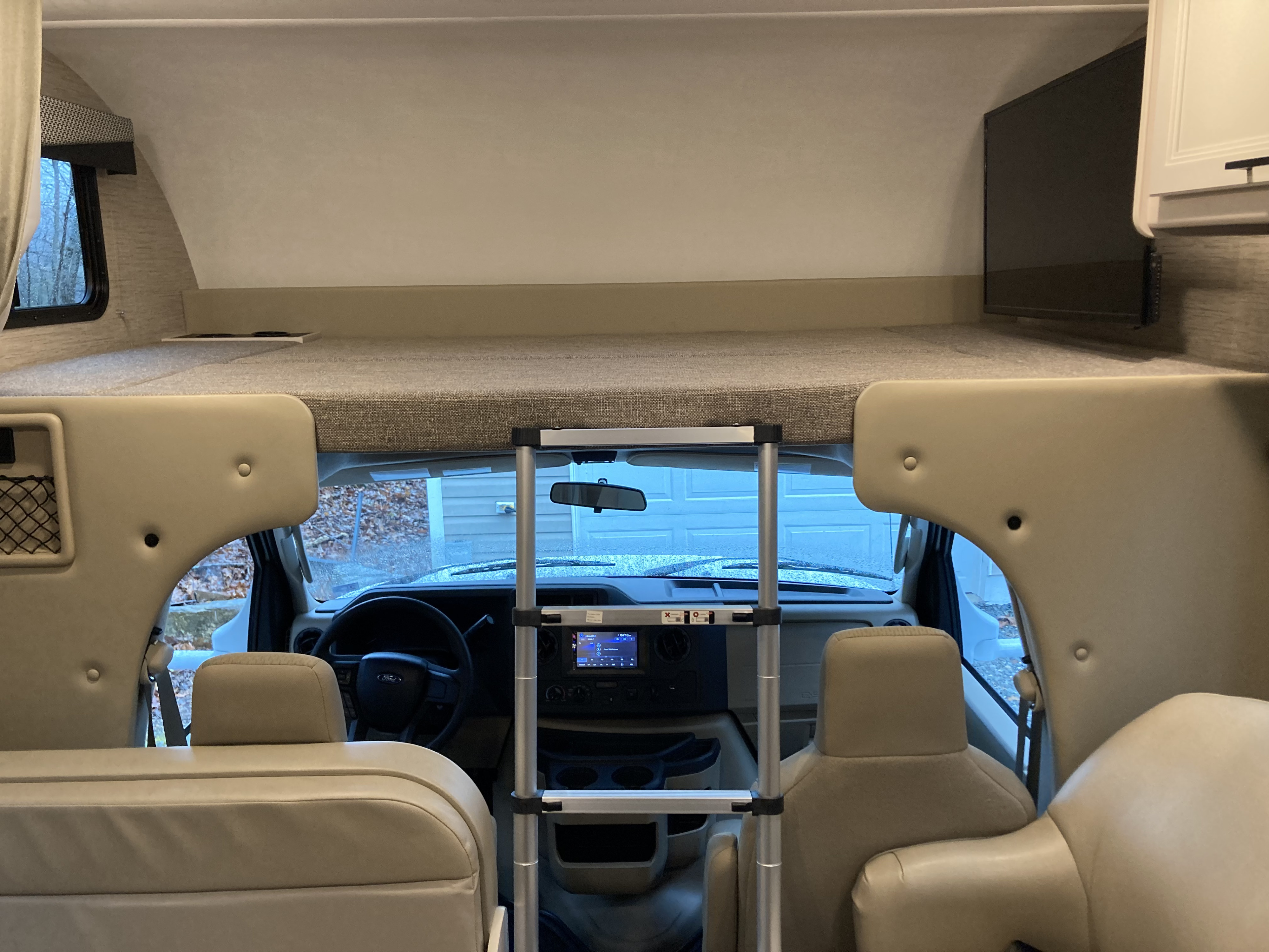 Interior view of an RV rental, featuring the over-cab sleeping bunk, access ladder, and the driver's area with dashboard and seats.. Thor Freedom Elite 2022