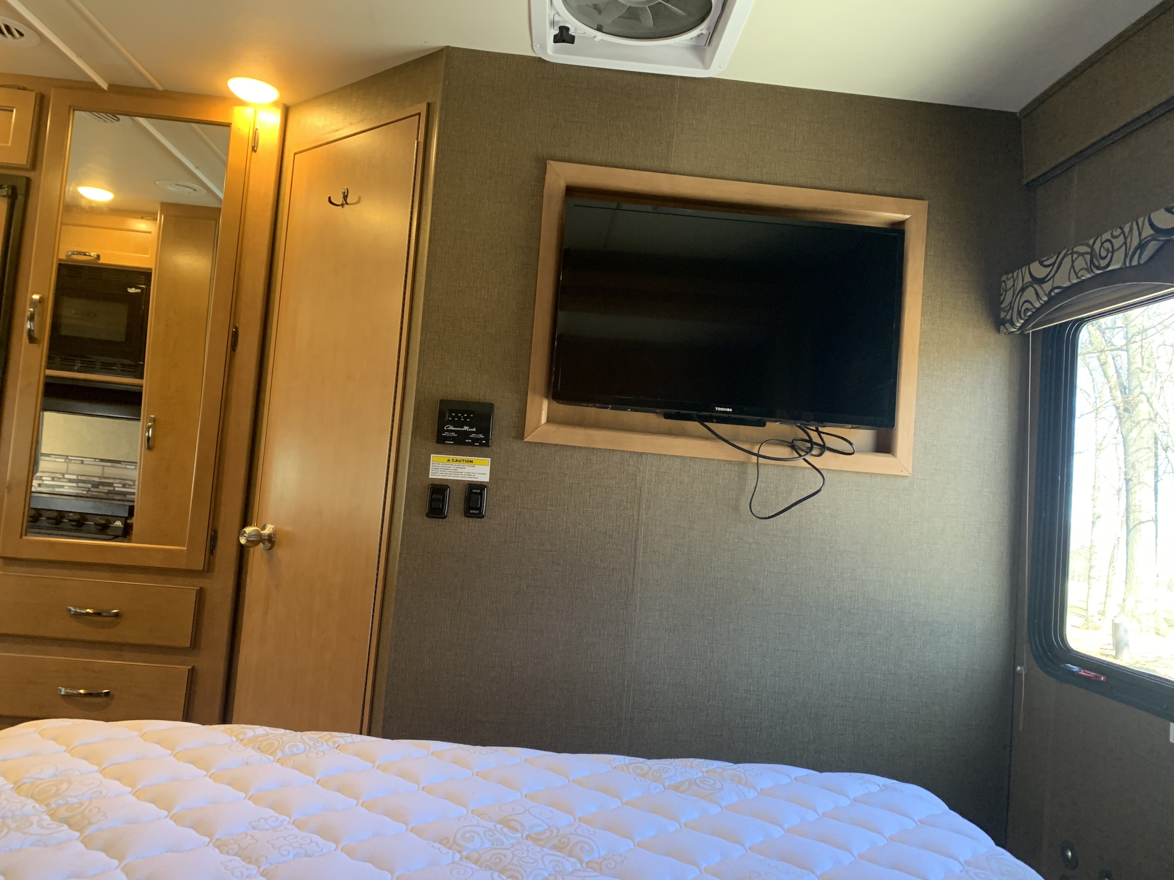 Interior of an RV rental, featuring a bed, mounted TV, reflected kitchen appliances, and window with trees outside.. Thor Four Winds 2017