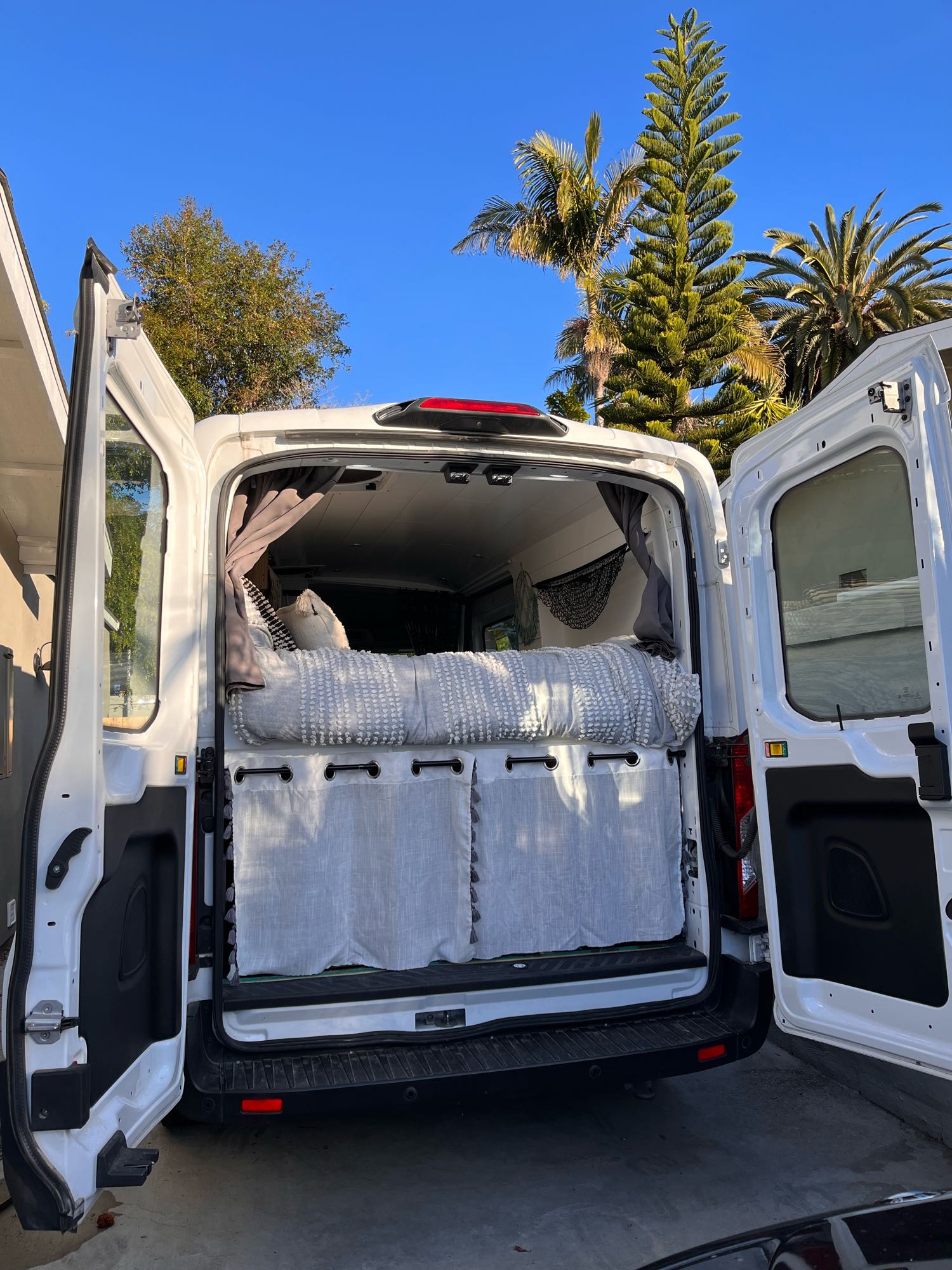 White camper van (rv rental) with open rear doors, showing a cozy bed, gray bedding, & privacy curtains inside. Sunny day, palm trees.. Ford Transit 2019