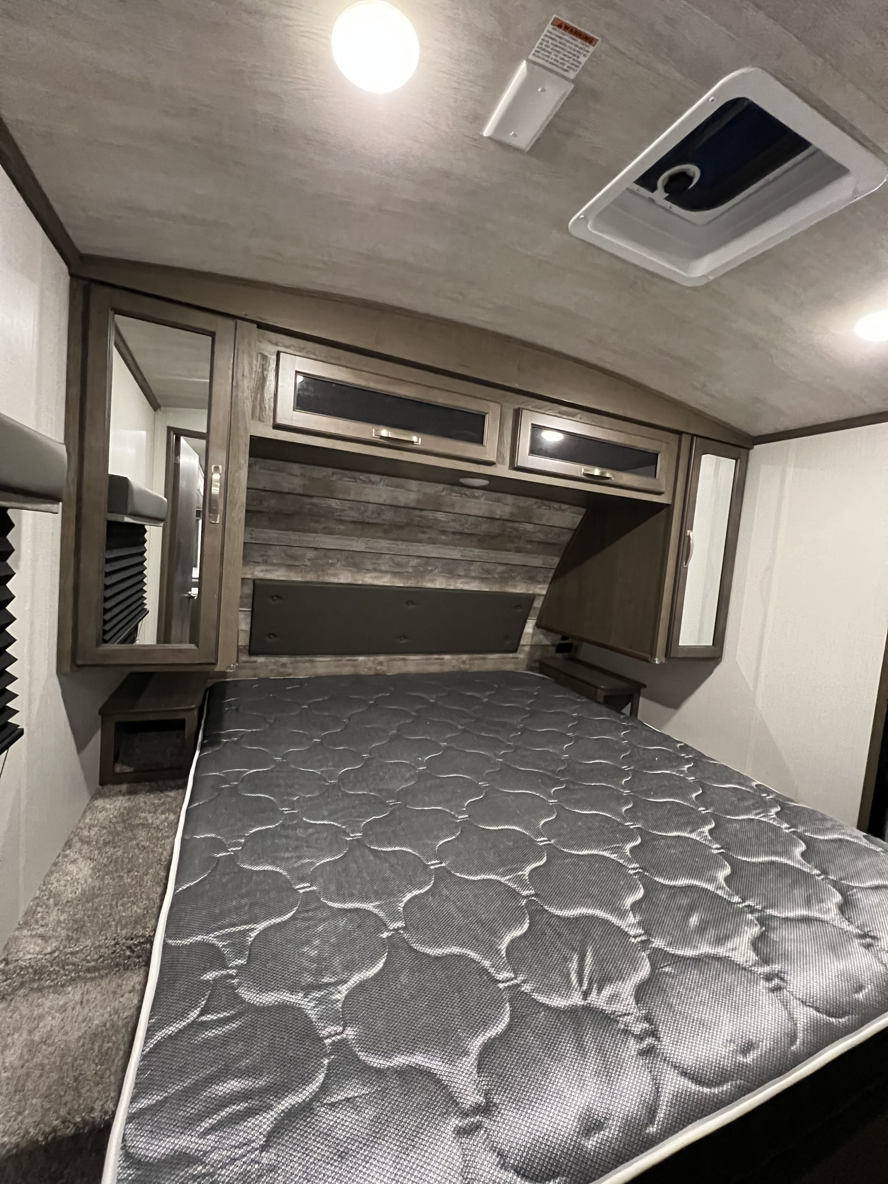 A cozy RV rental bedroom features a grey mattress, wood-paneled headboard, overhead cabinets, and a ceiling vent.. Keystone Cougar Half-Ton 2021