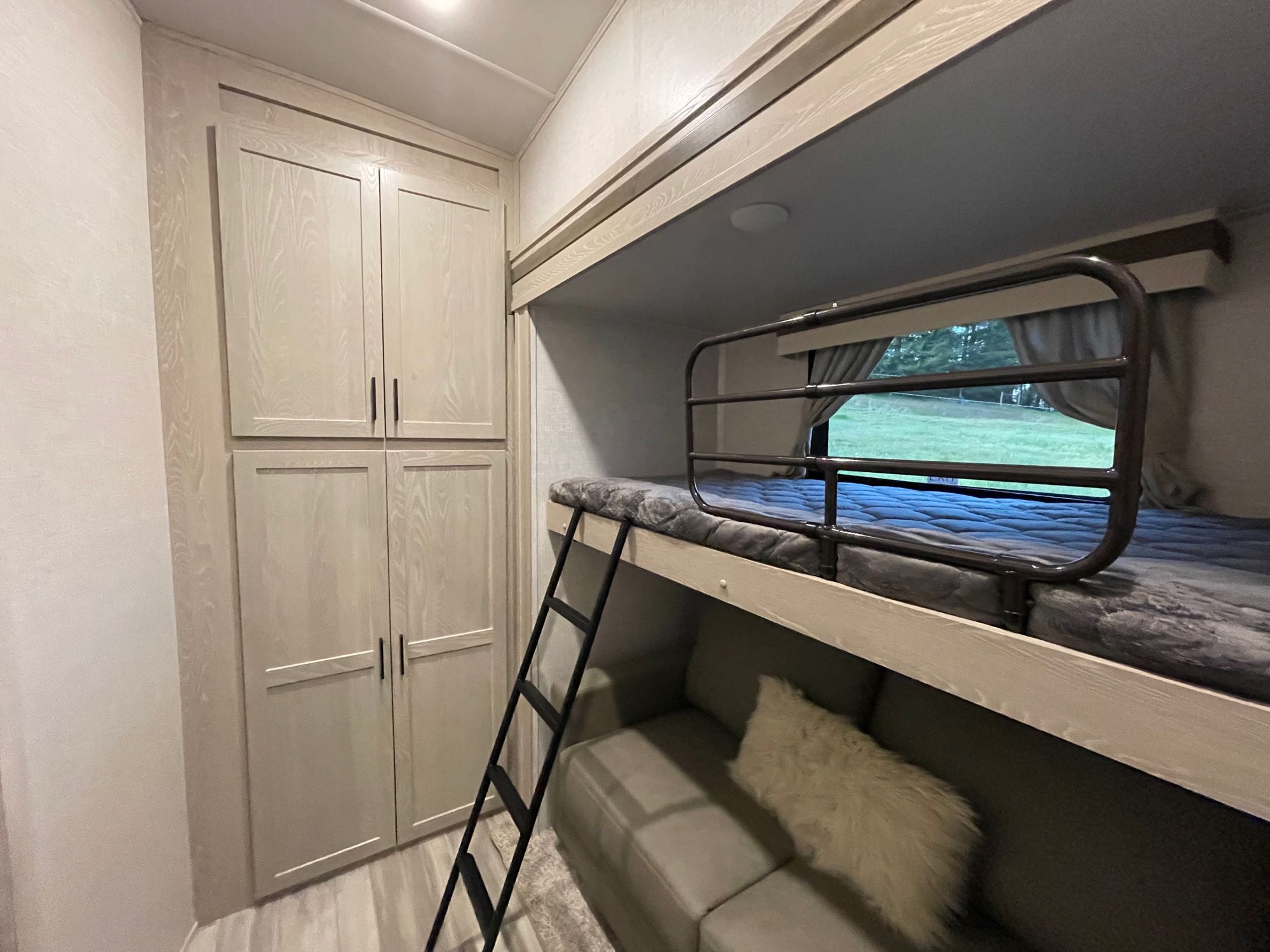 RV rental's bunk bed area features a top bunk with safety rail, lower sofa, ladder, and tall light cabinets. A window reveals outdoors.. Forest River Other 2022