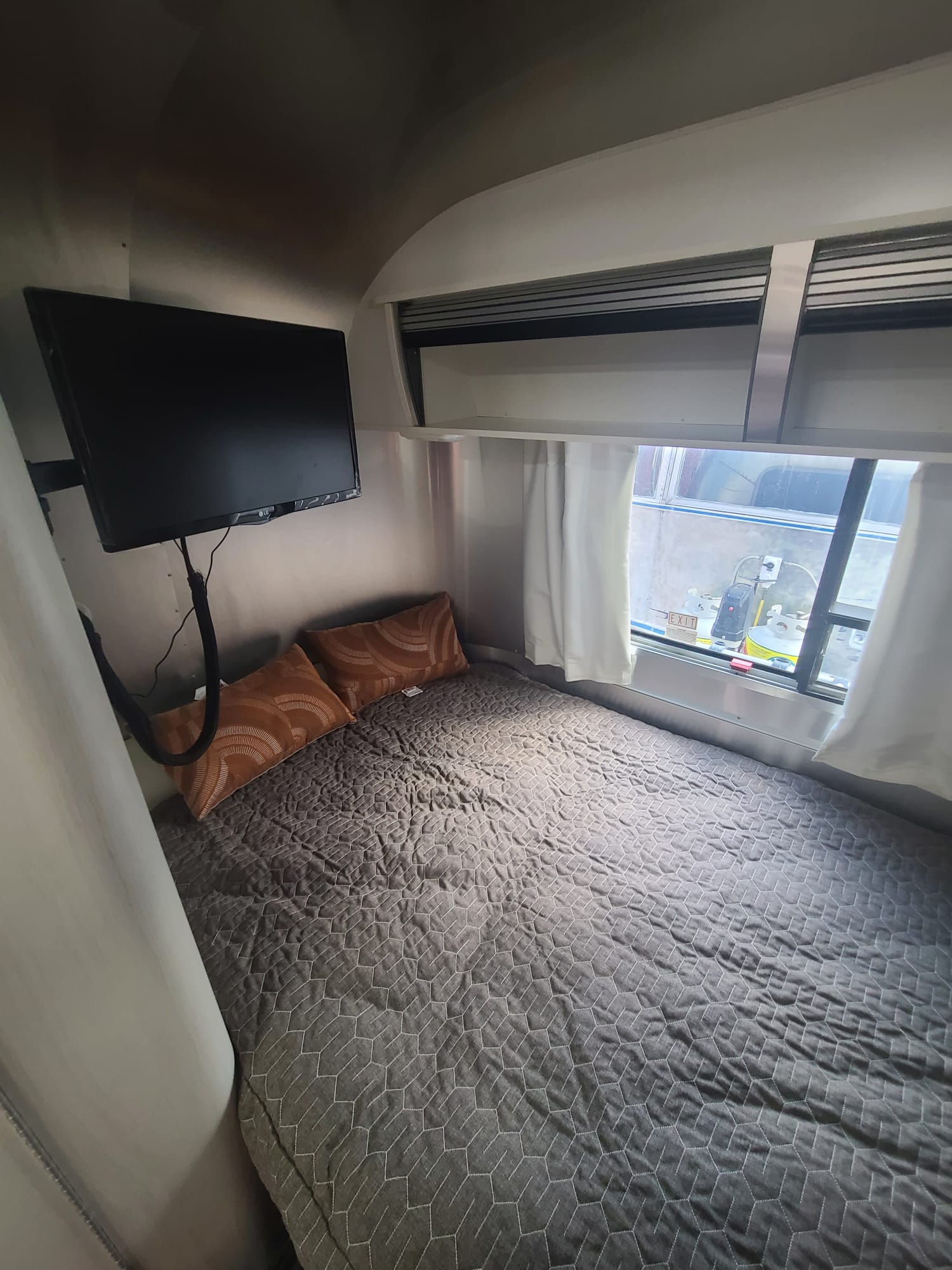 Cozy RV rental bedroom with a gray geometric quilt, orange pillows, and a TV mounted above the bed. Window shows exterior.. Airstream Sport 2017