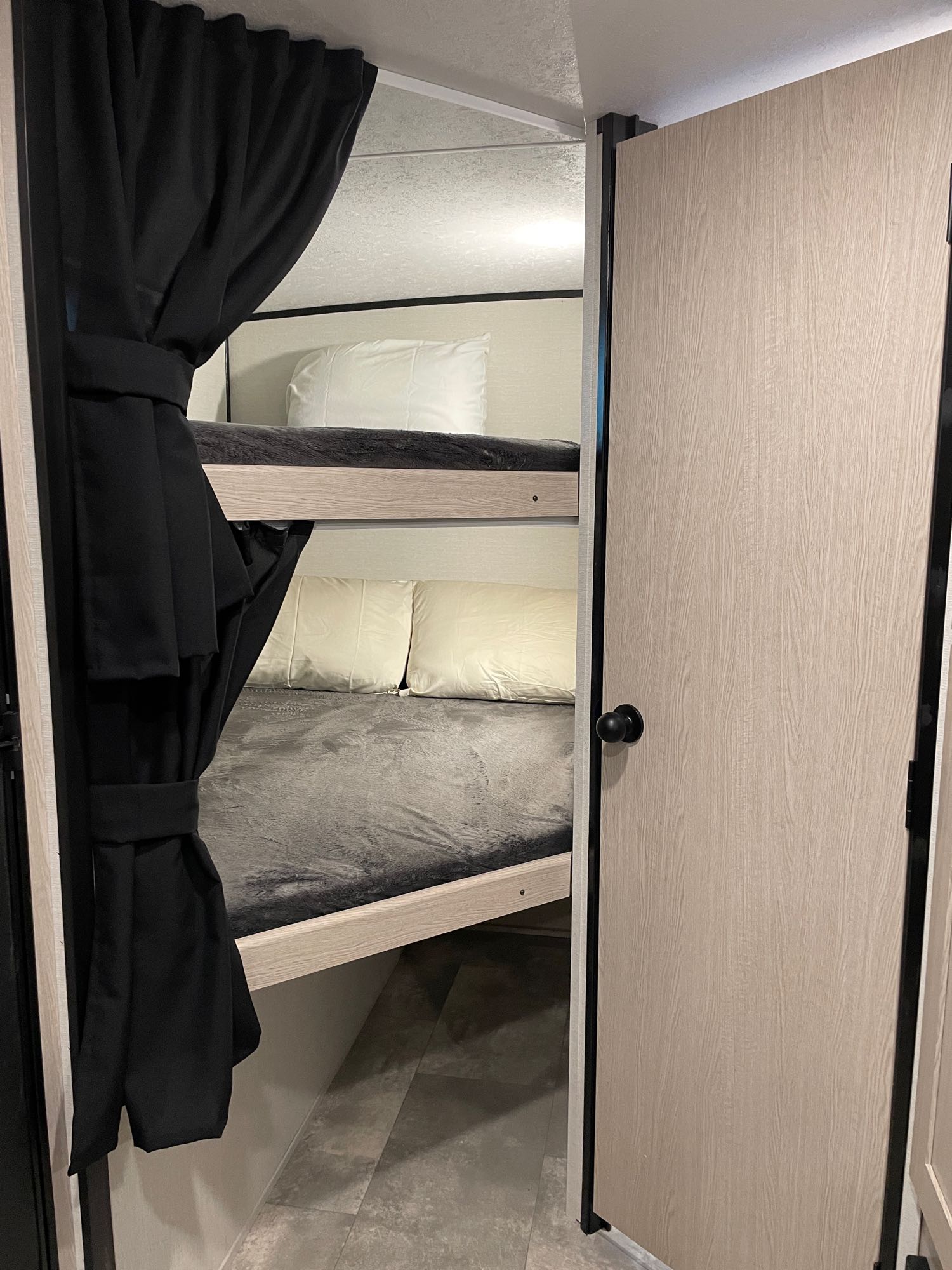 Inside an RV rental, cozy bunk beds with dark privacy curtains next to a light wooden door offer a compact sleeping area.. Coachmen Apex Nano 2022