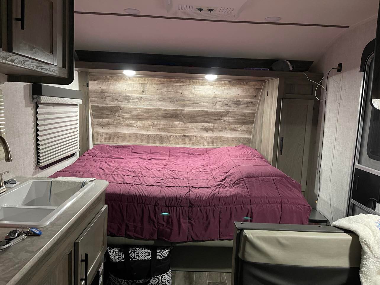 This RV rental features a cozy bedroom with a large purple bed, sink, cabinets, and window blinds, perfect for travel.. Gulf Stream Enlighten 2022
