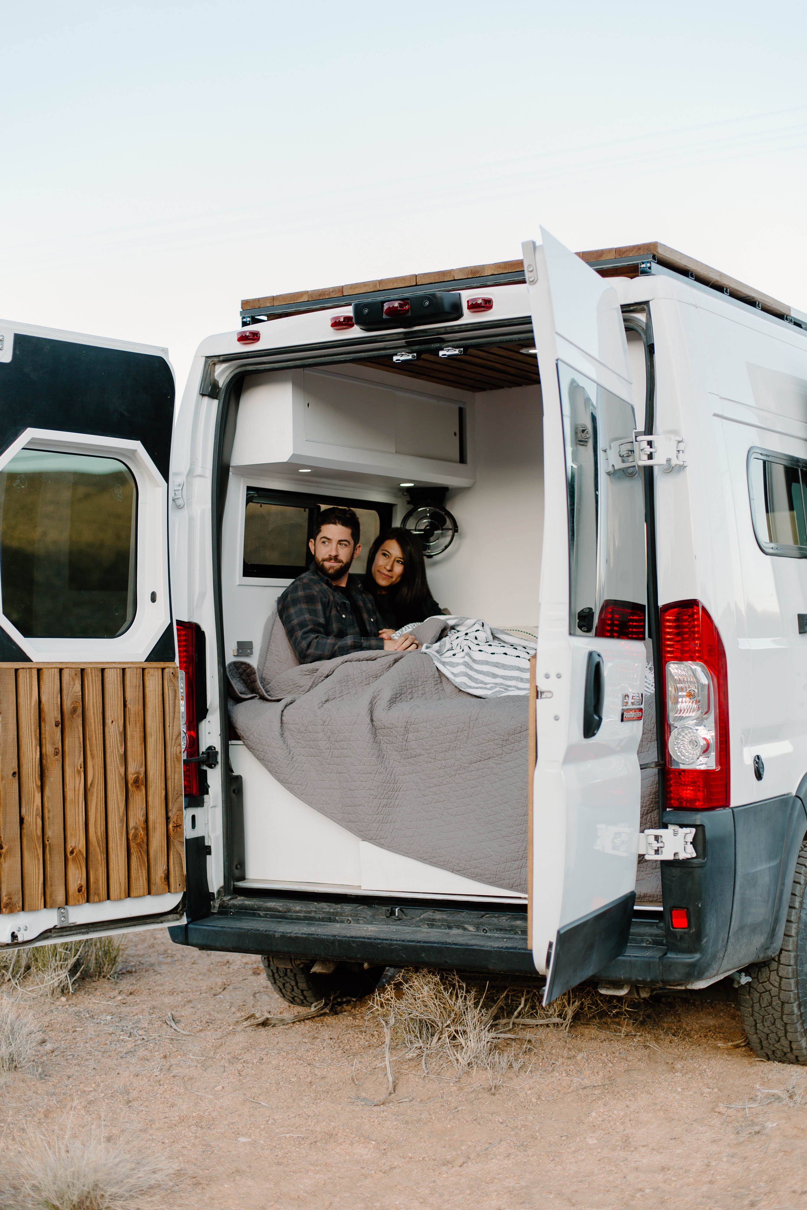 RV rental: A couple relaxes in their cozy camper van with open back doors, enjoying the desert view.. Dodge Sprinter 2019