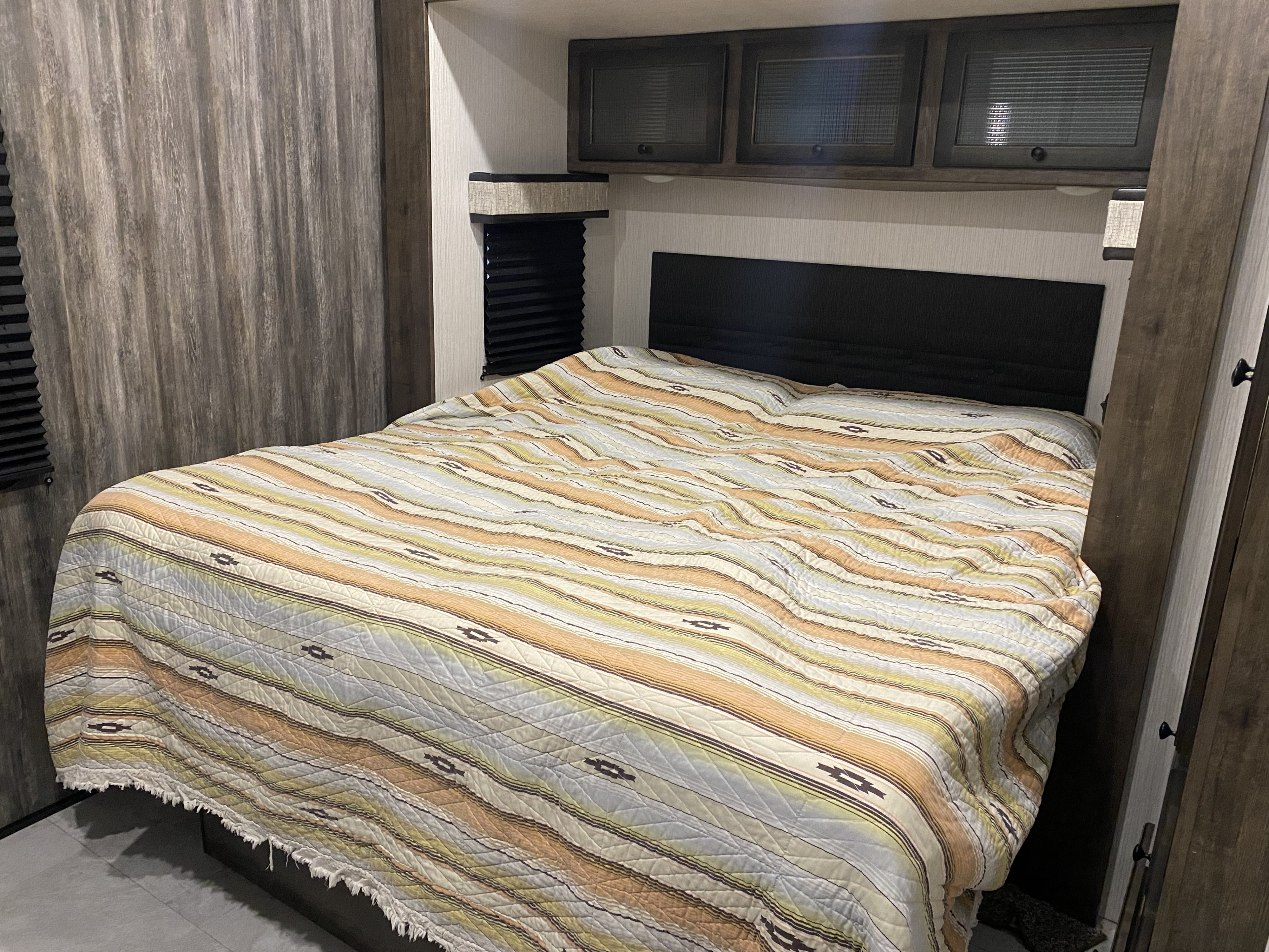 Cozy RV rental bedroom with a colorful striped bedspread, overhead cabinets, and wood-paneled walls. Perfect for travel comfort.. Heartland Milestone 2021