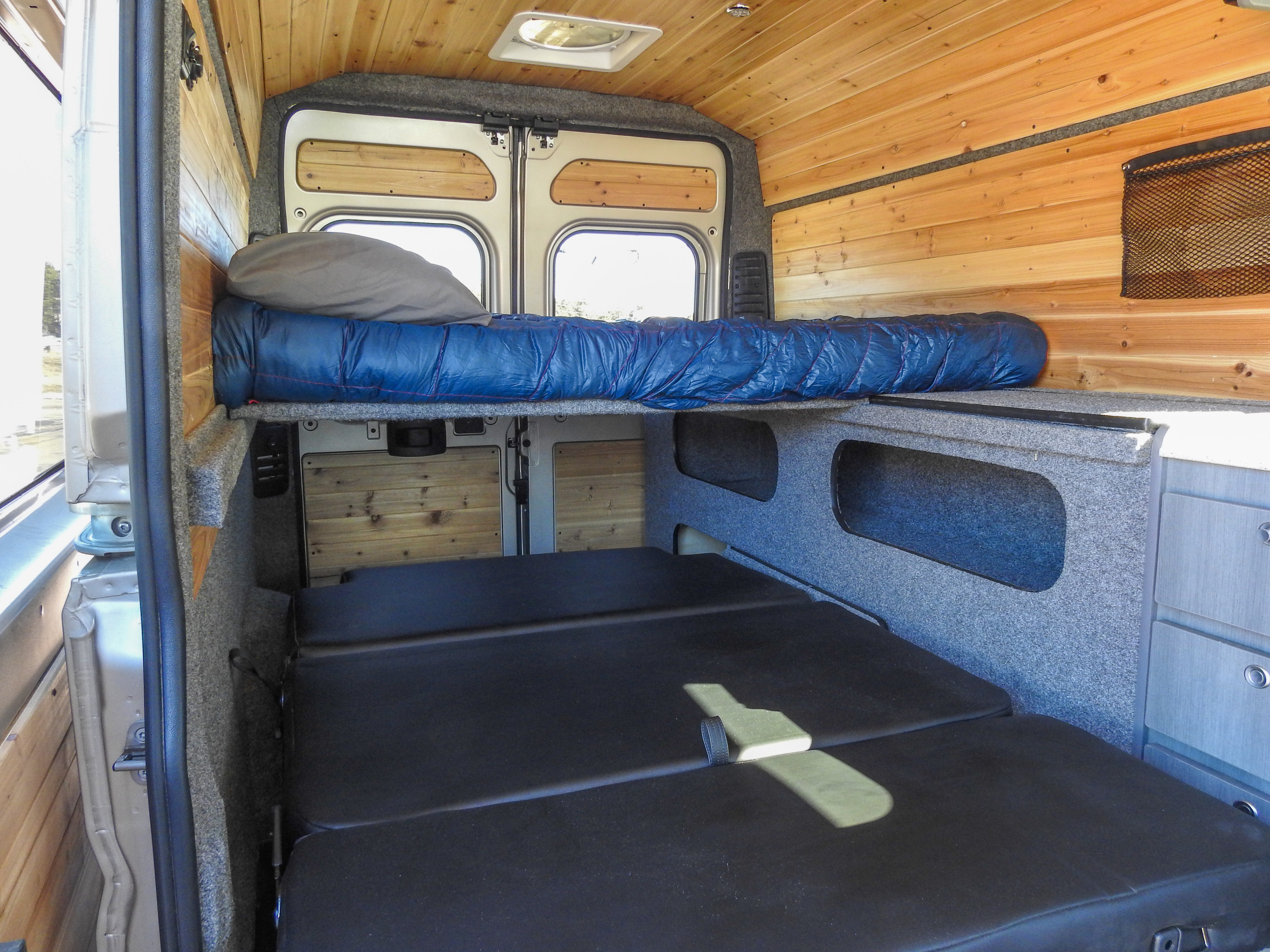 Inside a modern RV rental: a wood-paneled camper van with a cozy bed, lower seating/bed, and smart storage.. Dodge Promaster- Summit X Edition OA9 2021