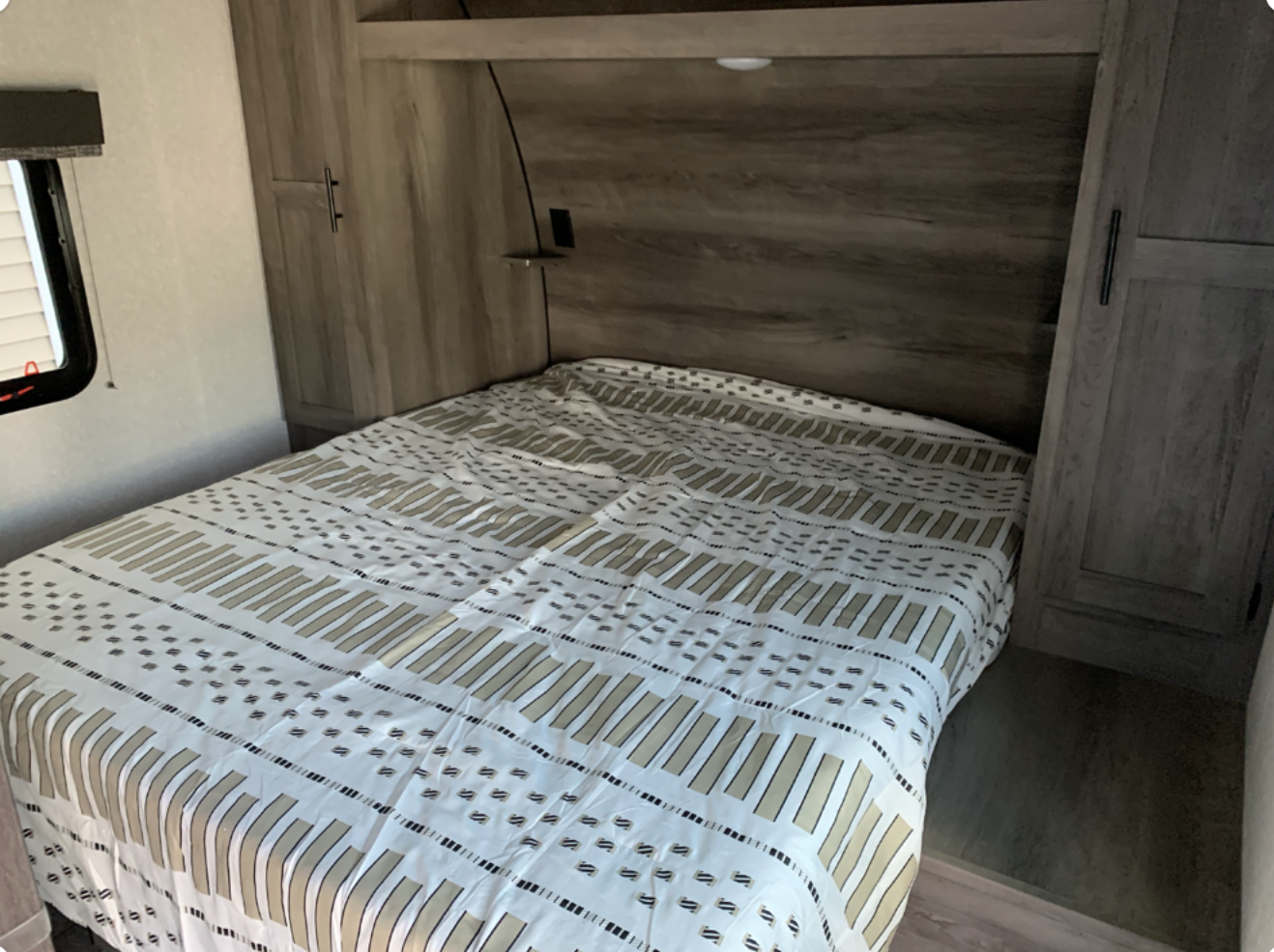 Comfortable RV rental bedroom with a full bed, patterned duvet, wood cabinets, and a window with blinds.. kz sportsman 2021