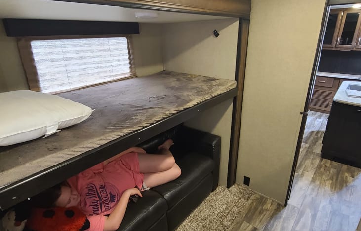 An RV rental interior shows stacked bunk beds; a person relaxes on the lower bunk's couch, kitchen beyond.. GRAND DESIGN 312BHTS 2019