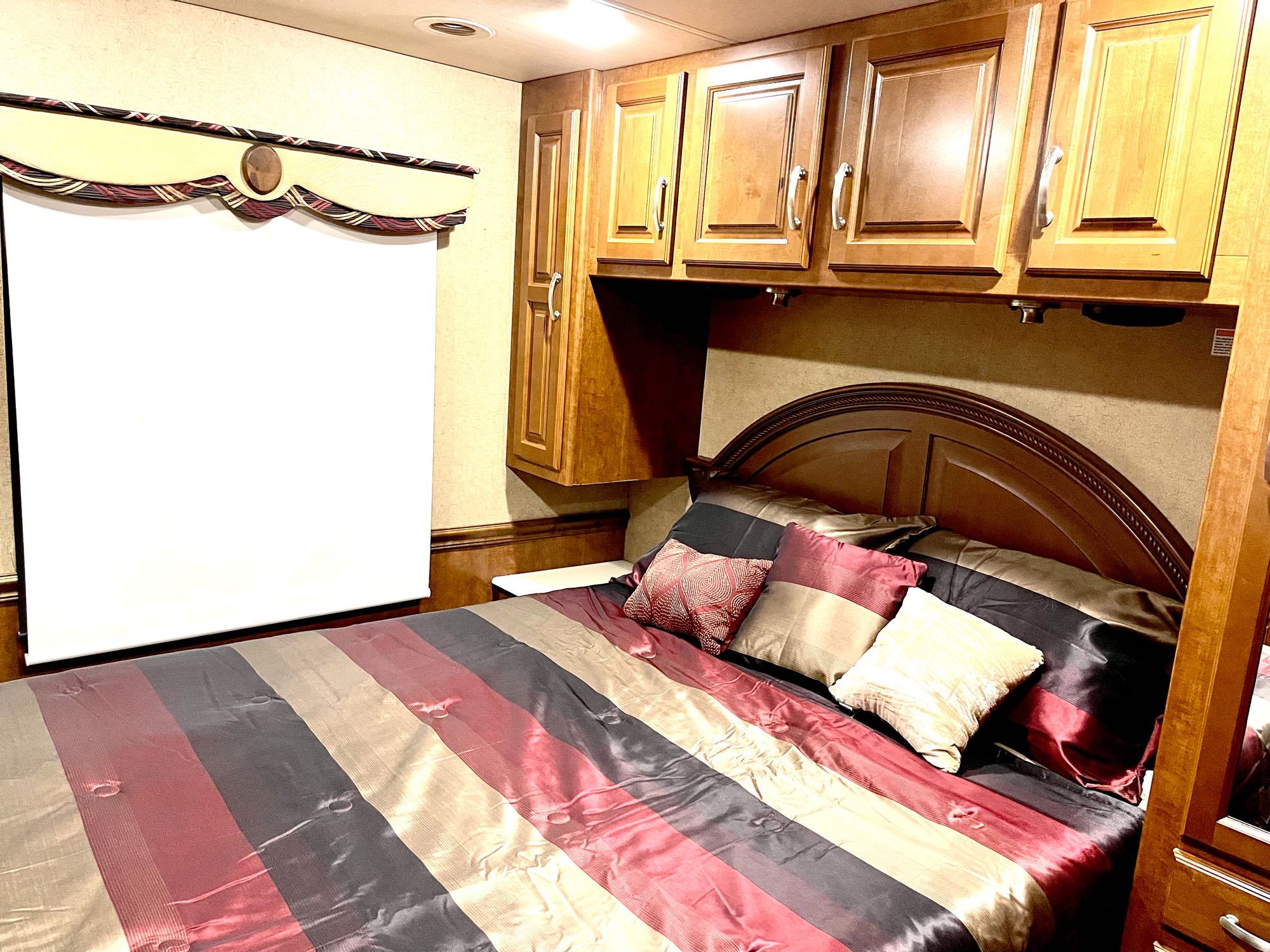 This inviting RV rental bedroom features a striped bed (red, black, tan), wood cabinets, and a window, ensuring a comfortable stay.. Thor Challenger 2015