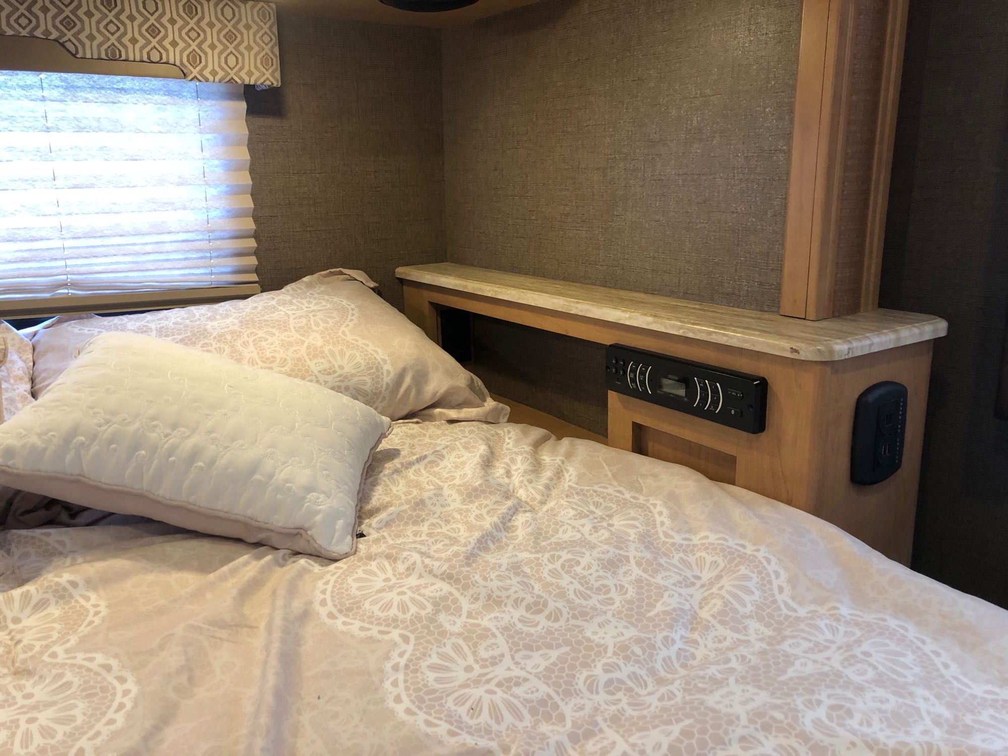 Cozy RV rental bedroom features a bed with elegant lace-patterned bedding, integrated controls, and window blinds.. Thor Vegas 2017