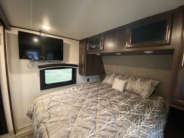 Inside an RV rental bedroom, featuring a comfy bed with patterned comforter, mounted TV, window, and overhead storage.. Jayco Jay Flight 2020