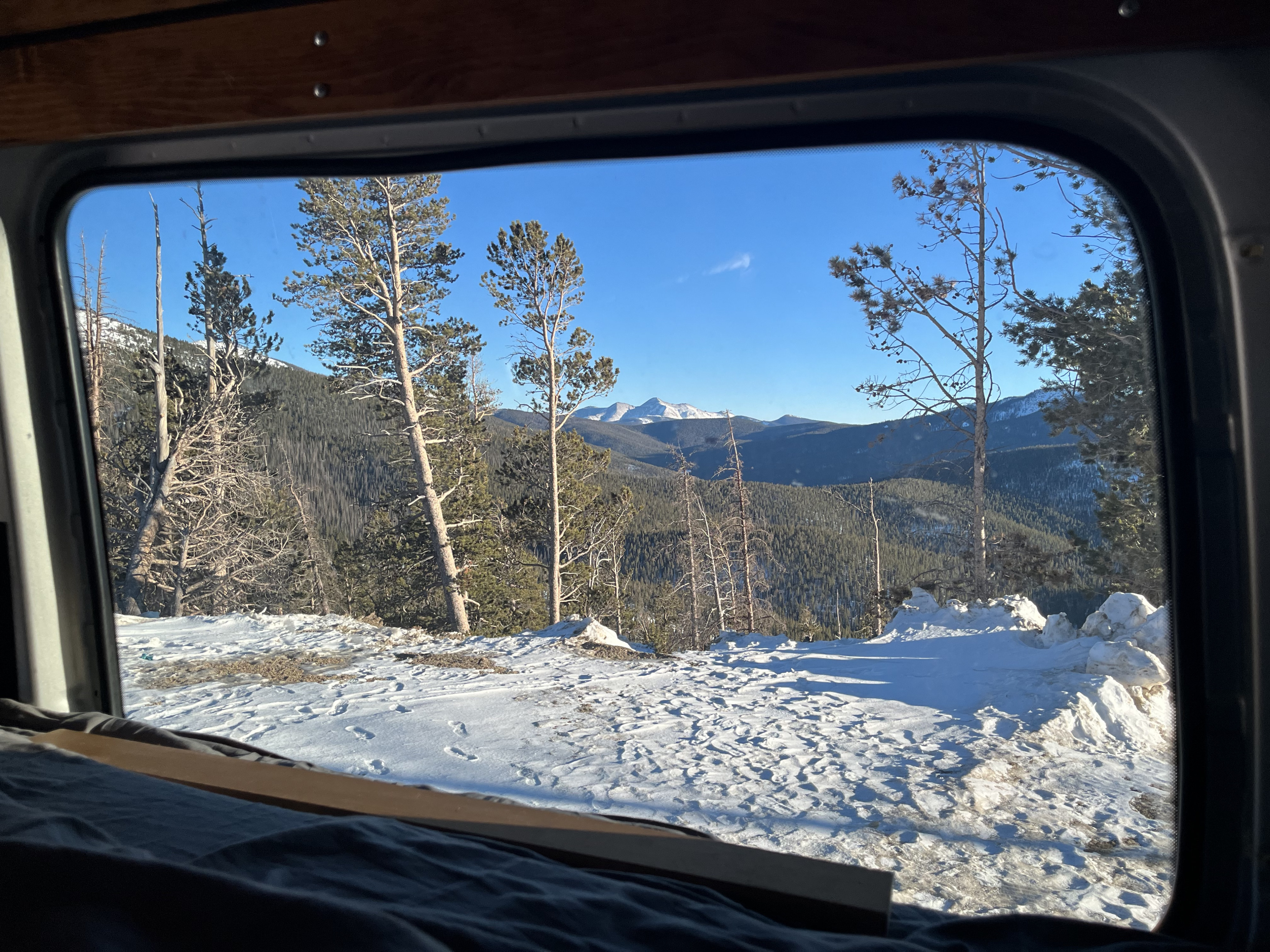 View from cozy RV rental bed, overlooking snowy mountains and pine trees. A serene winter landscape unfolds outside.. Mercedes-Benz Sprinter 2014
