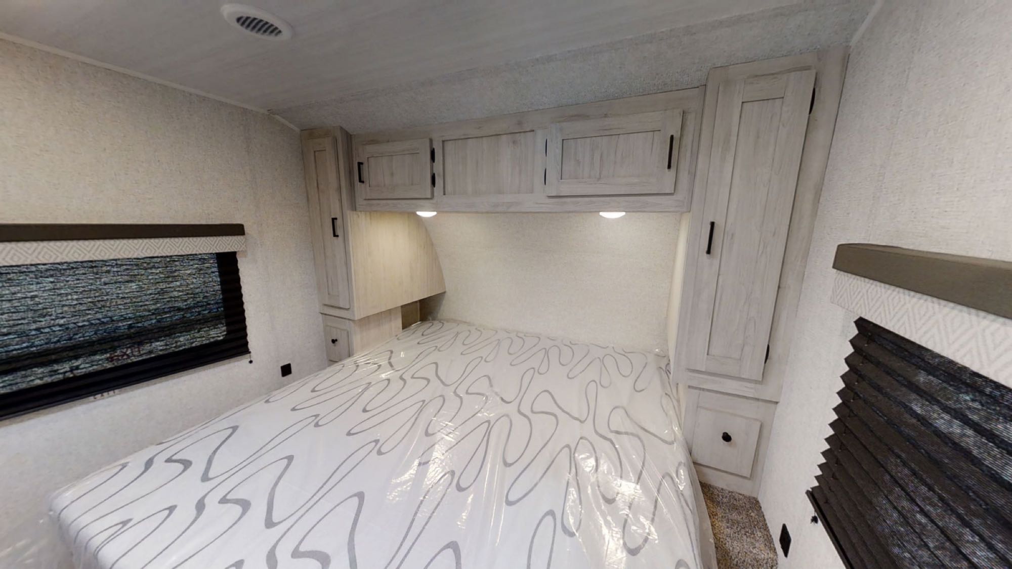 RV rental bedroom featuring a patterned bed, wood-tone overhead cabinets, and side windows with blinds.. East to West Della Terra 2022