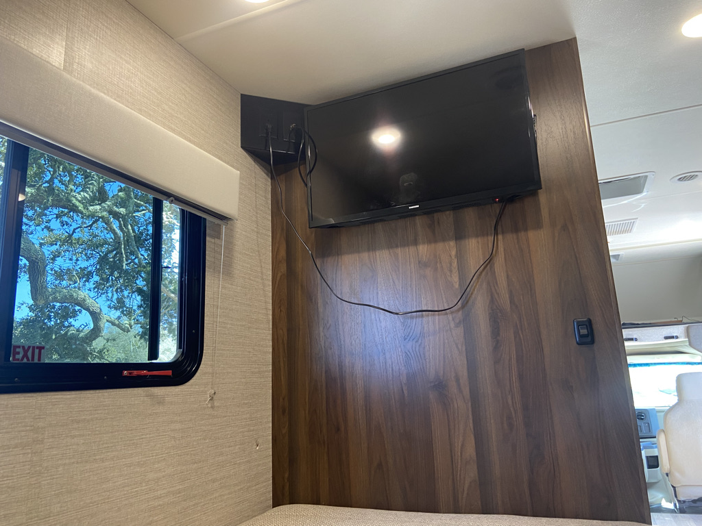 Inside an RV rental: TV mounted on a rich wood wall, with a window and pull-down shade.. Winnebago Outlook 2020