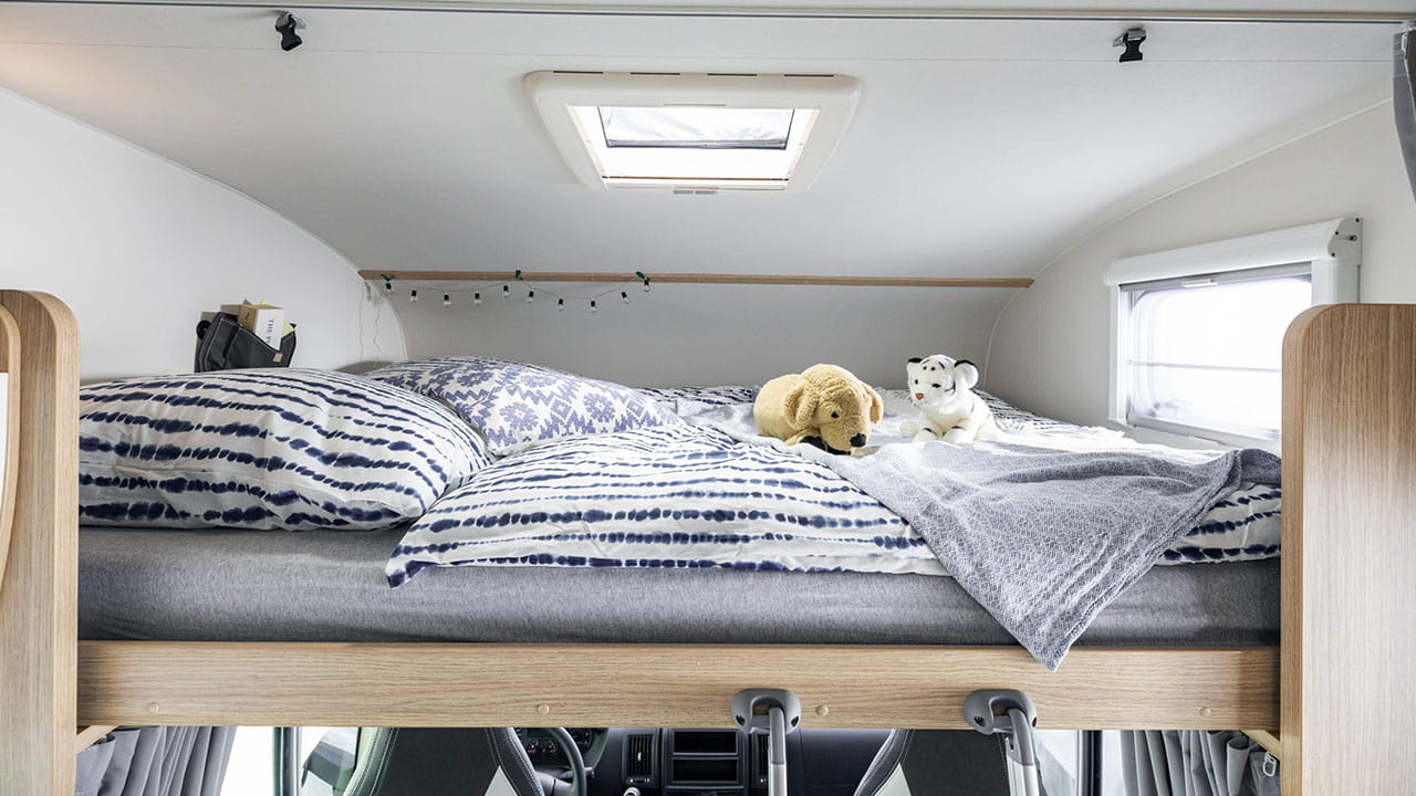 Inside an RV rental: a bright, cozy cab-over bed with blue-and-white bedding, pillows, and plush toys.. Sunlight A60 2022