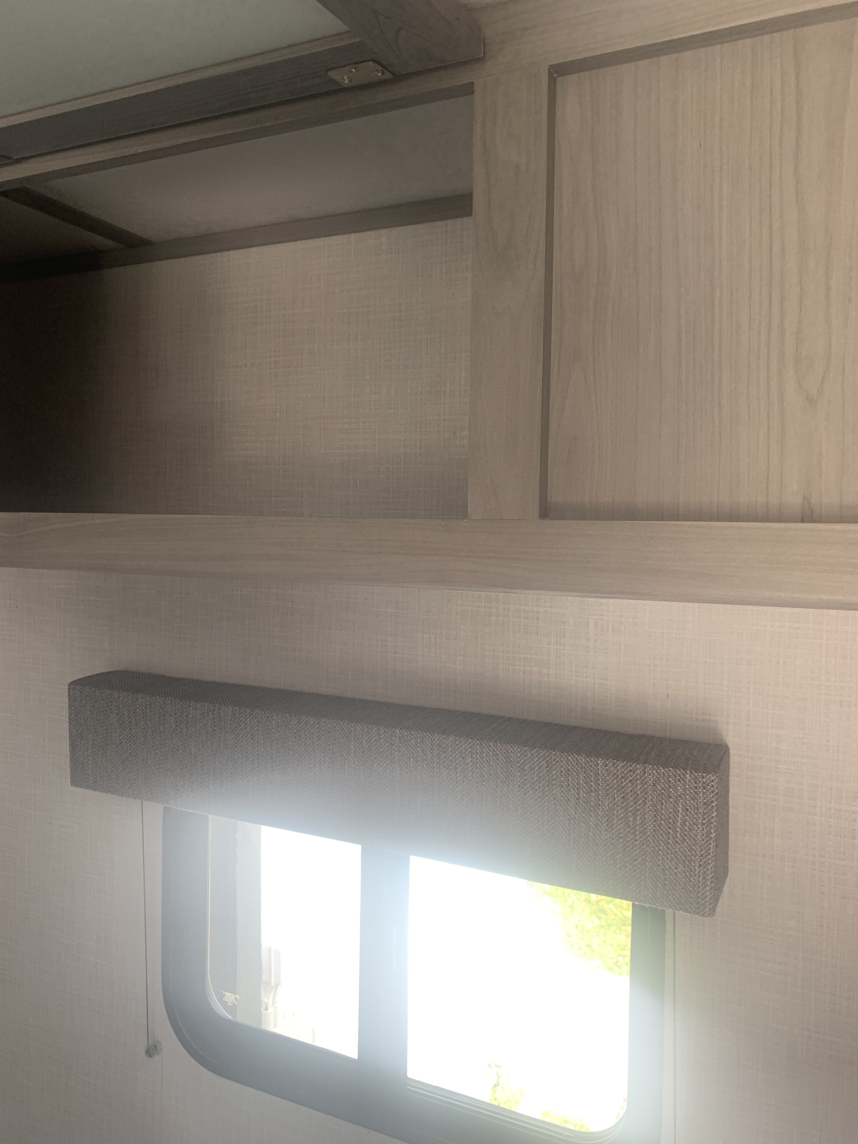 RV rental interior with light wood cabinets, shelves, and a window covered by a gray fabric shade. Bright light enters.. Coleman 17B 2022