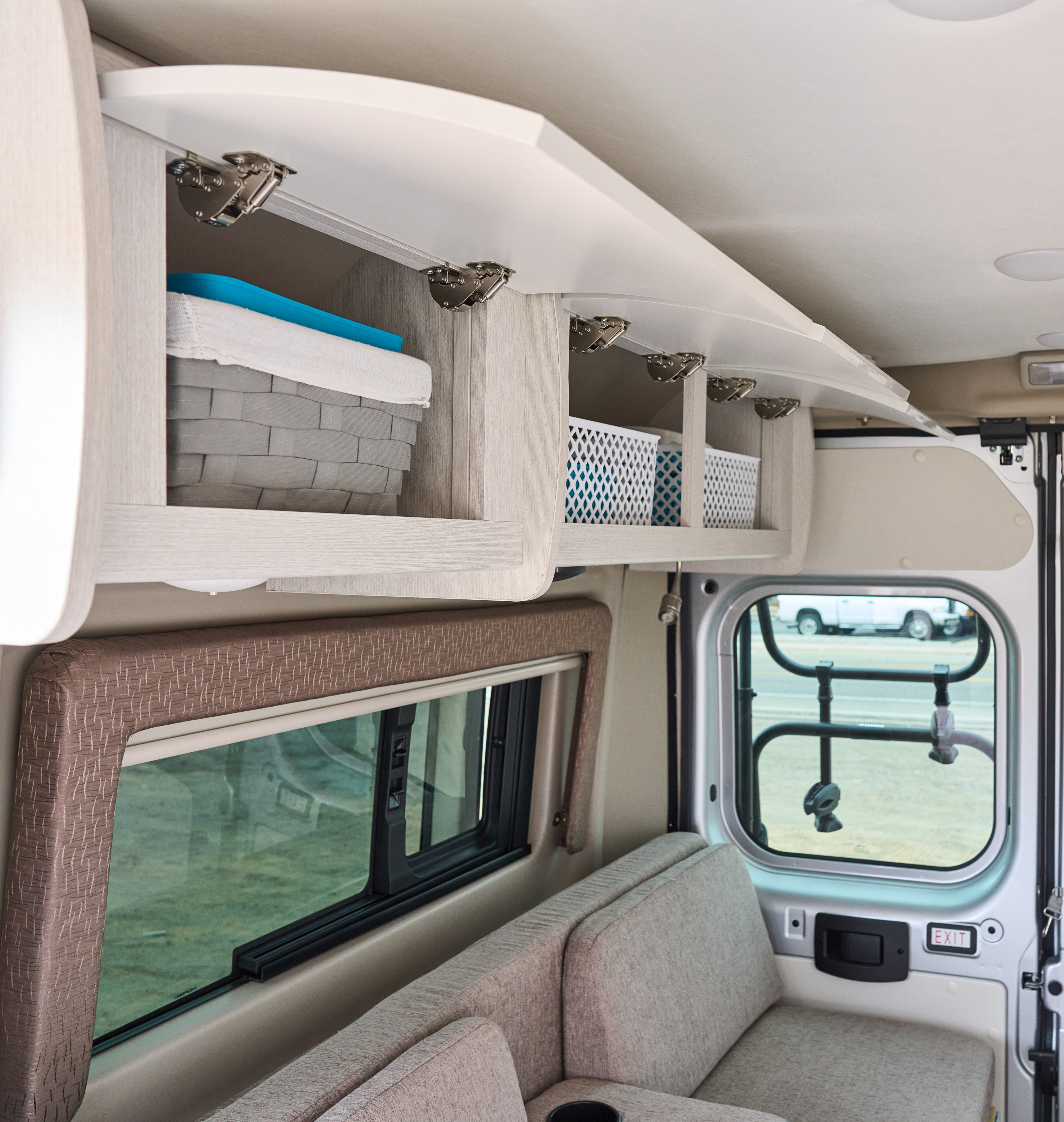 Interior of an RV rental showing open overhead cabinets with storage baskets, comfortable seating, and a window view.. Thor Rize 2022