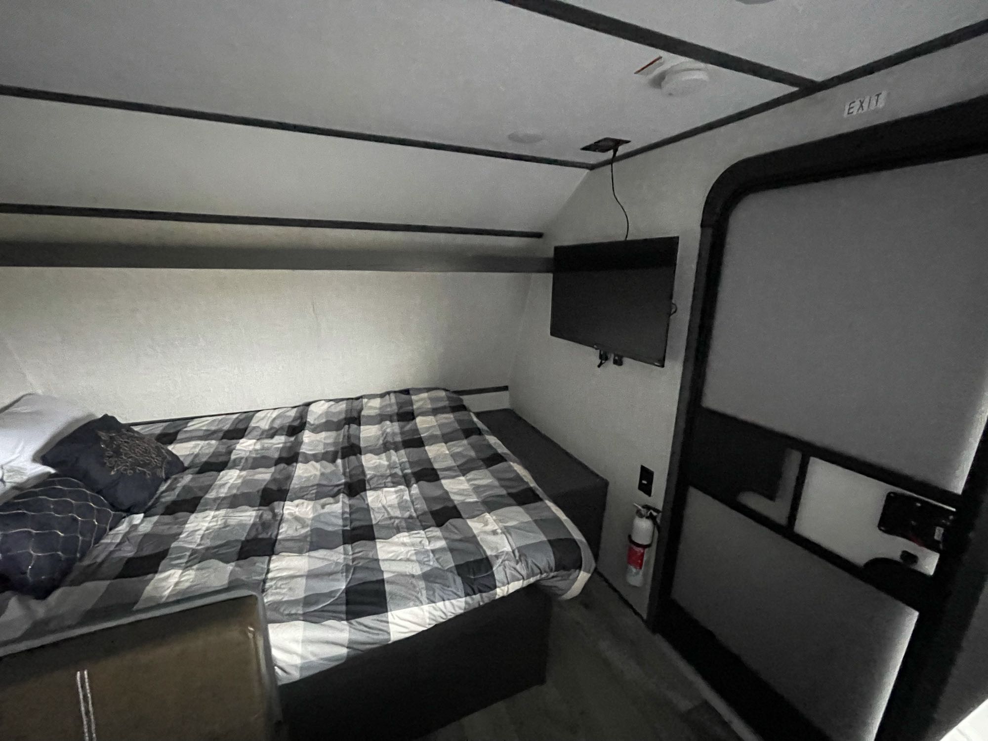 Cozy RV rental bedroom featuring a plaid bed, wall-mounted TV, and an exit door with a fire extinguisher.. Dutchmen Rv Aspen Trail 17BH 2022