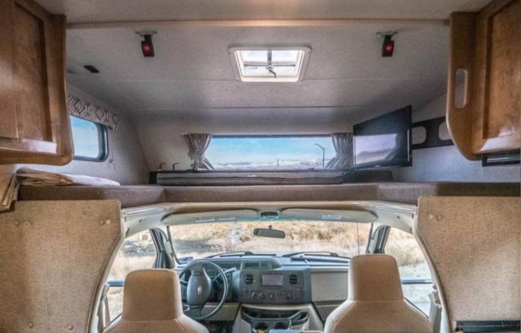 RV rental interior view: cab-over bunk with TV, driver's cockpit, and scenic window/skylight views. Ready for adventure!. Forest River Coachmen Leprechaun 2020