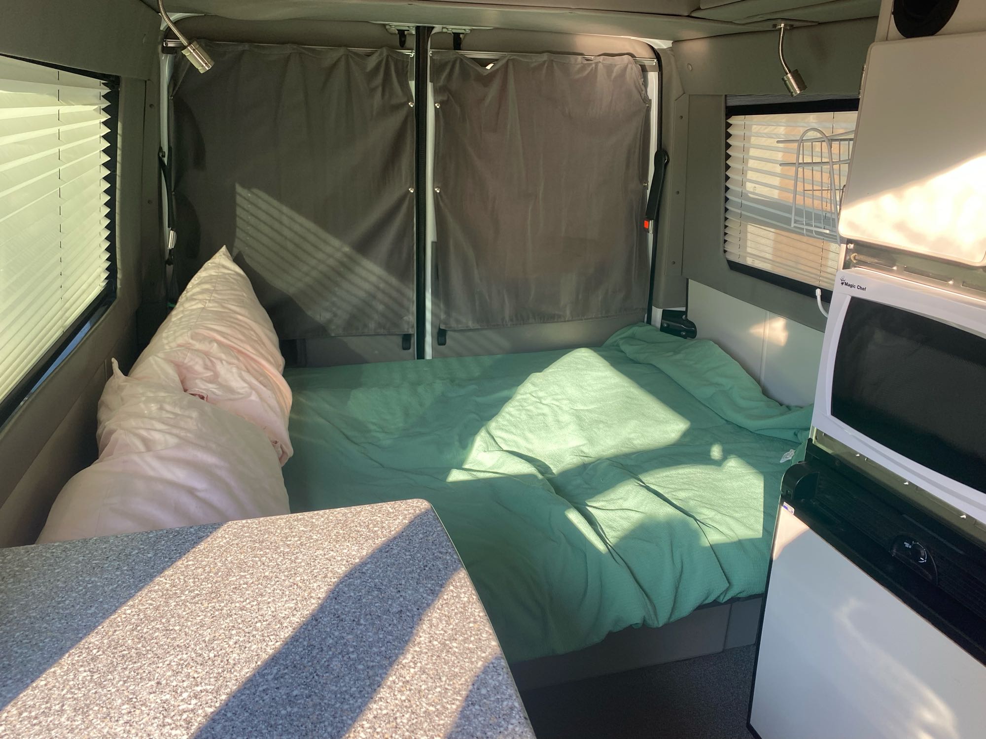An RV rental's inviting interior: bed with green bedding, window with blinds, and compact kitchen for comfort on the road.. Sportsmobile Mercedes Sprinter 2500 2012