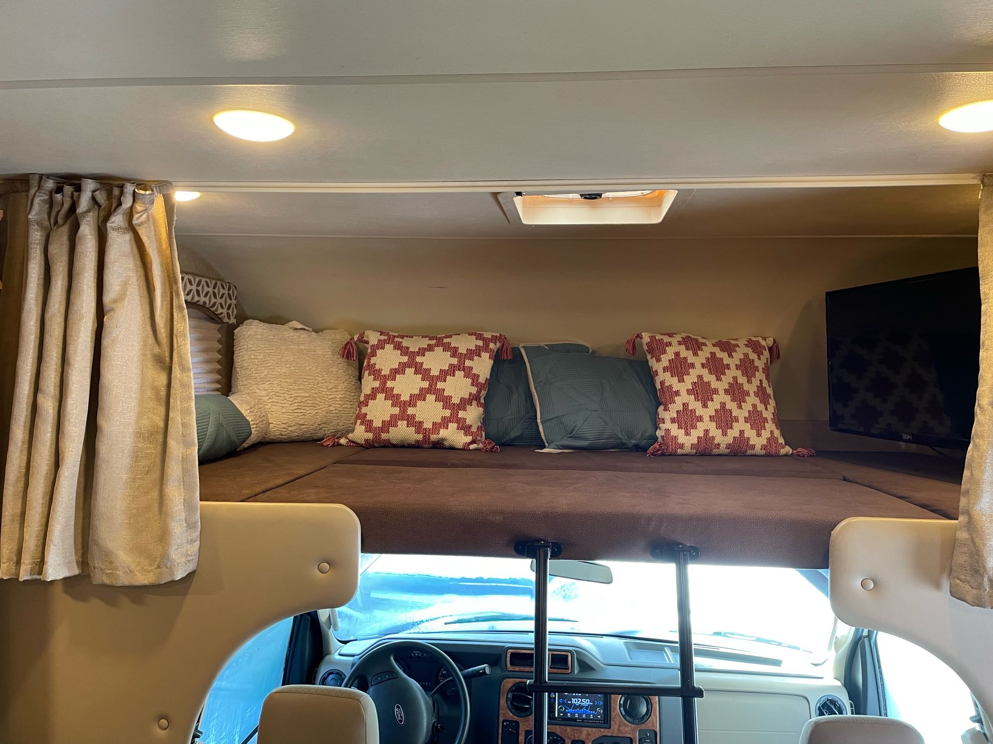 Interior of an RV rental, featuring the cozy cab-over bunk bed with pillows, curtains, and a TV, overlooking the cockpit.. Thor Four Winds 2017
