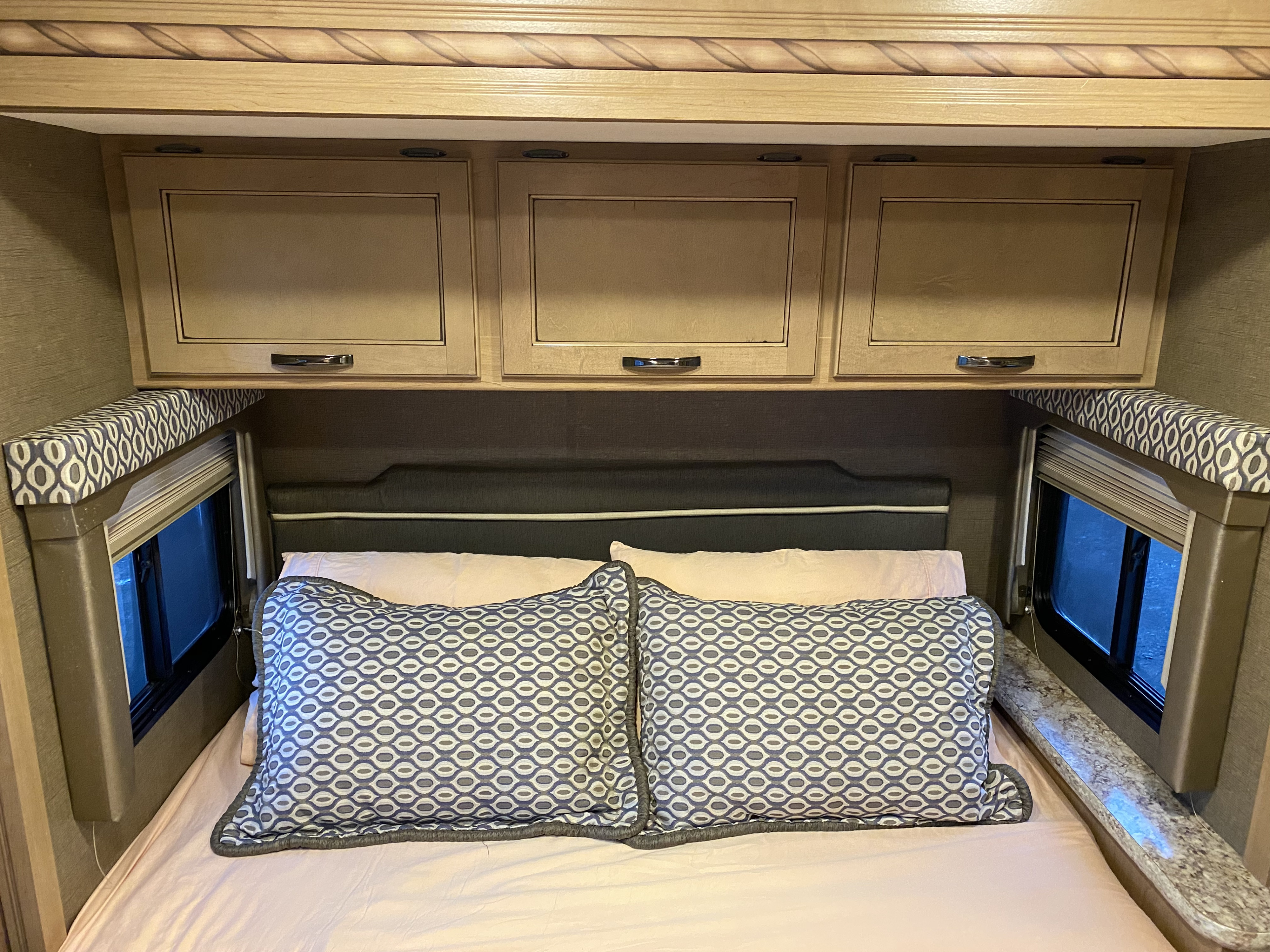 An RV rental bedroom featuring a comfortable bed, light wood overhead storage, and windows with patterned valances.. Thor Freedom Elite 2017