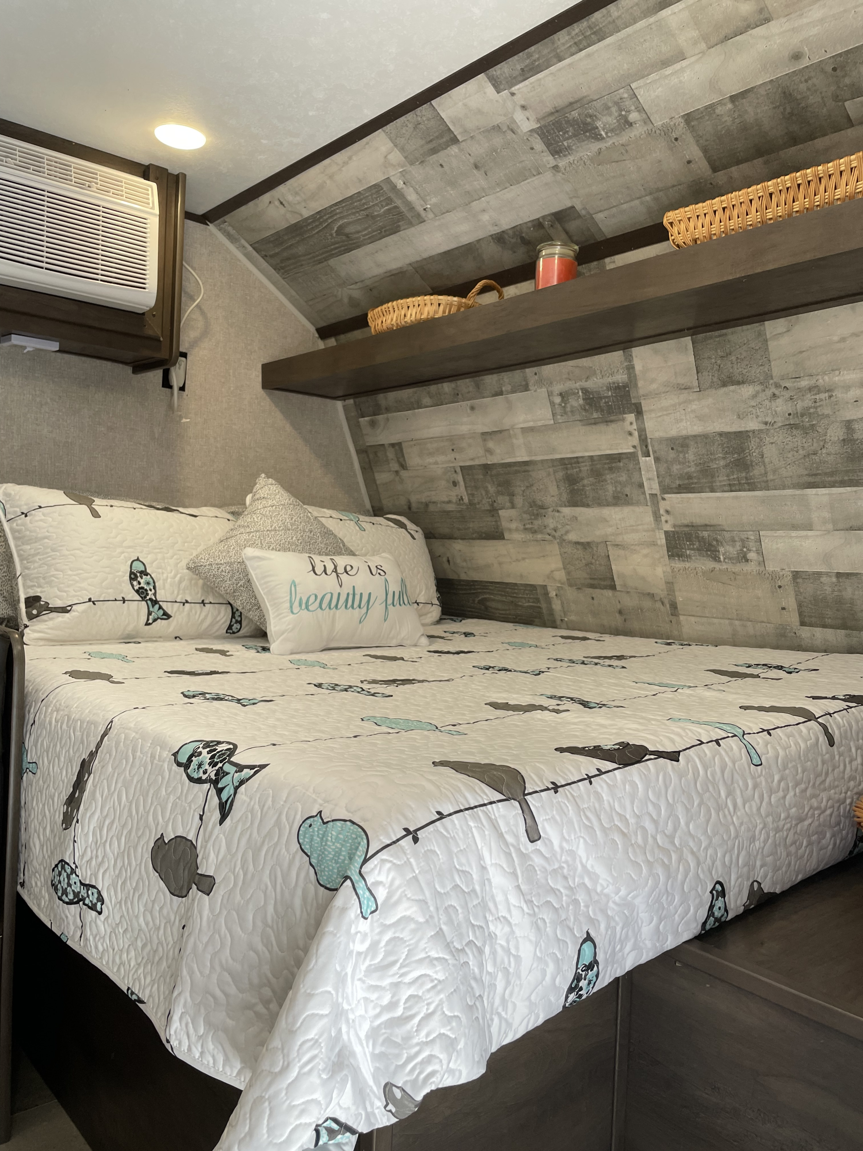 Inviting RV rental sleeping area, showcasing a comfortable bed with bird quilt, wall-mounted AC, and decor shelves.. Dutchmen Coleman Lantern 2022