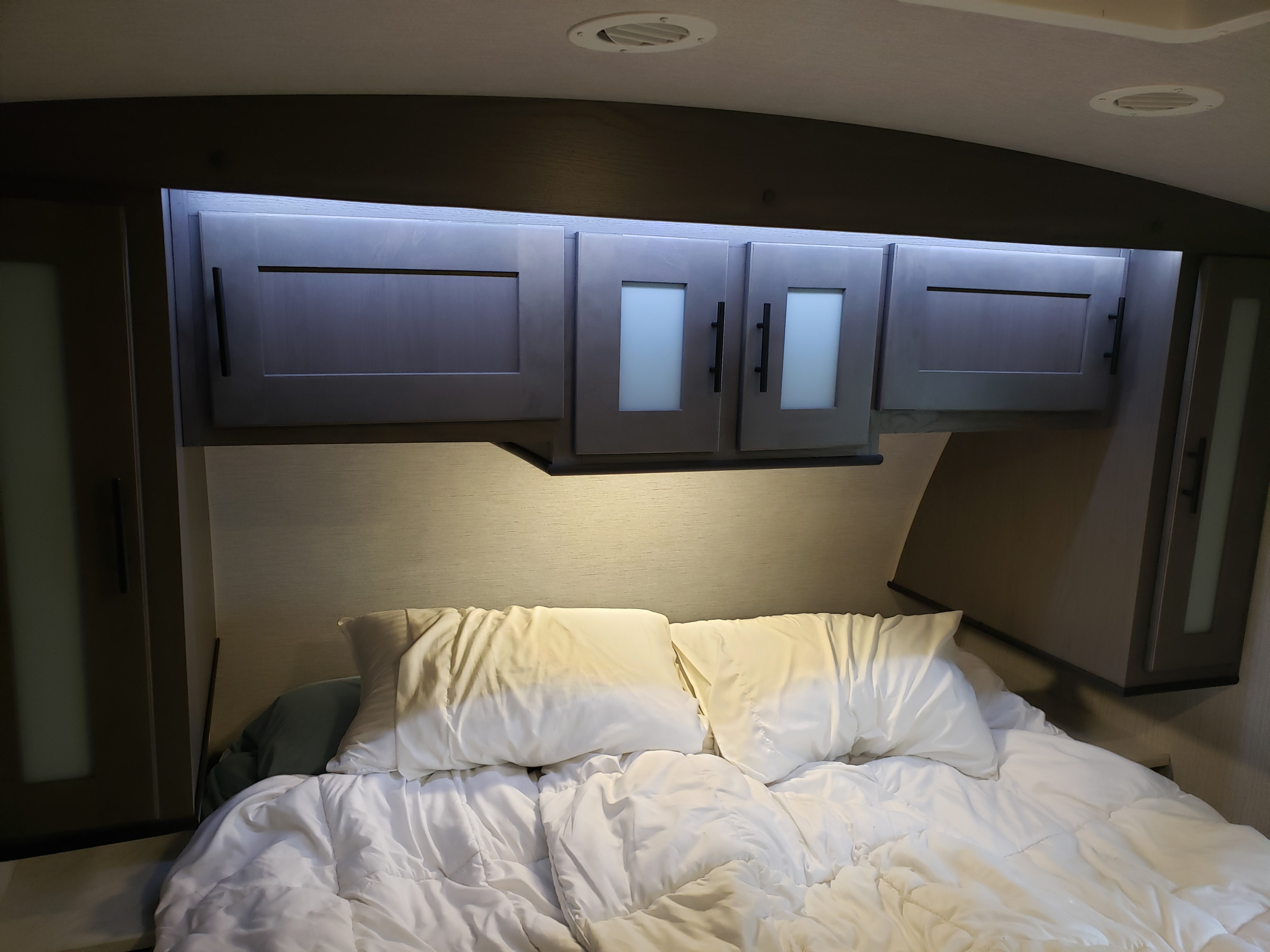 This RV rental features a compact bedroom with a bed, white pillows, and practical overhead storage with illuminated cabinets.. Cruiser RV MPG Ultra Lite 2020