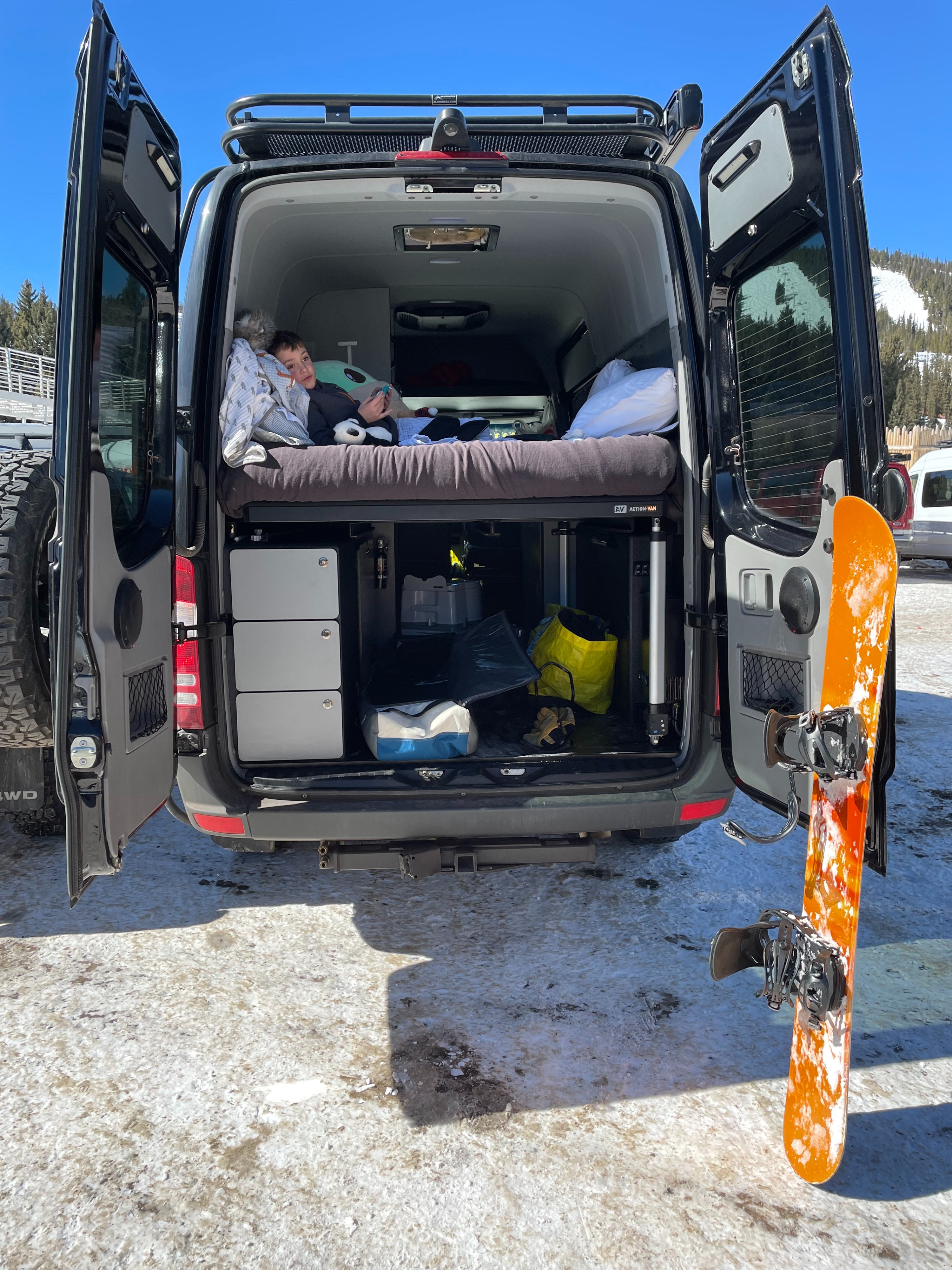 Cozy RV rental for winter trips! A child relaxes inside the camper van, with a snowboard ready for mountain slopes.. Sprinter Action Van 2016