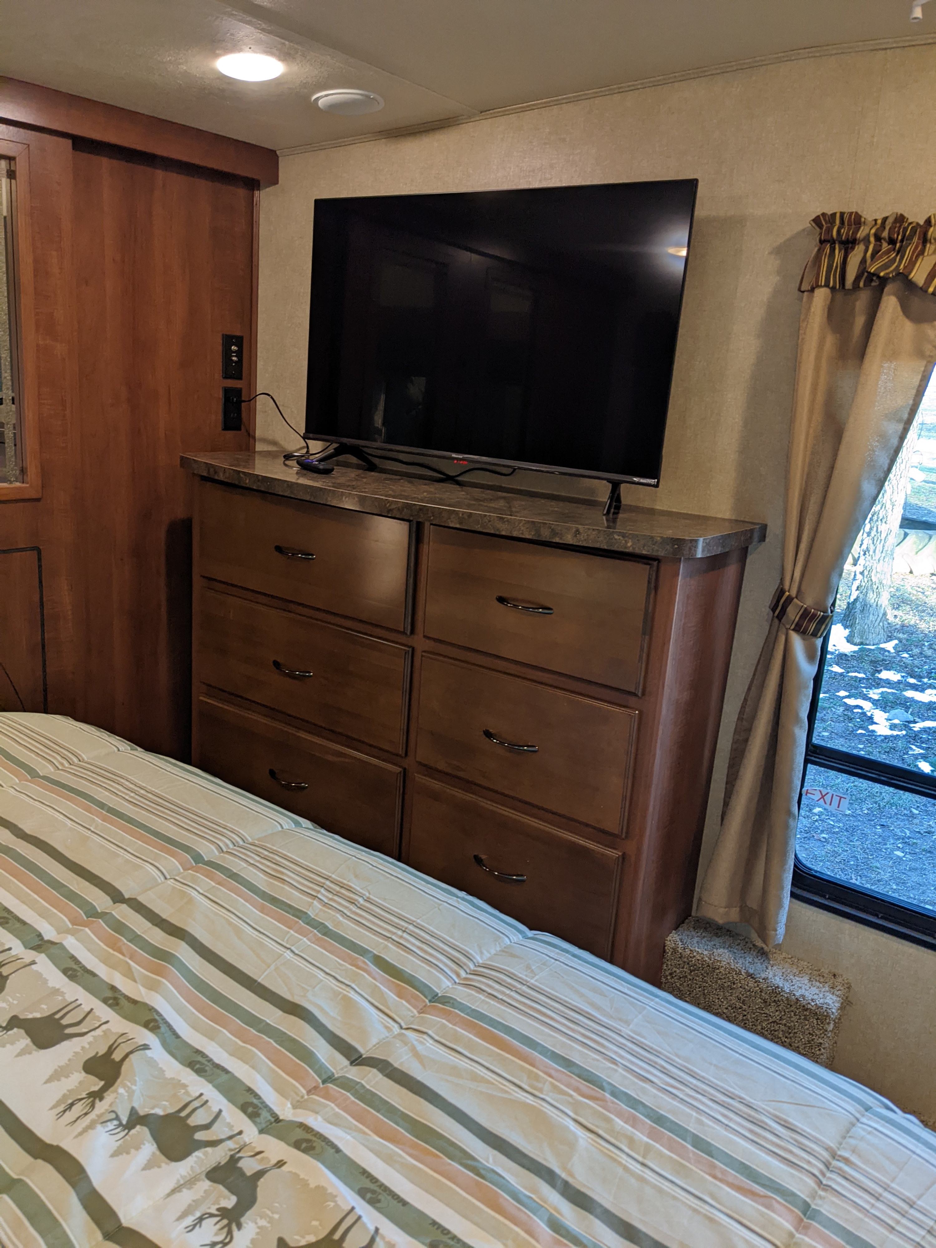 Inside an RV rental bedroom: bed with striped comforter, dresser, TV, and window view with winter landscape.. Forest River Sandpiper 2015
