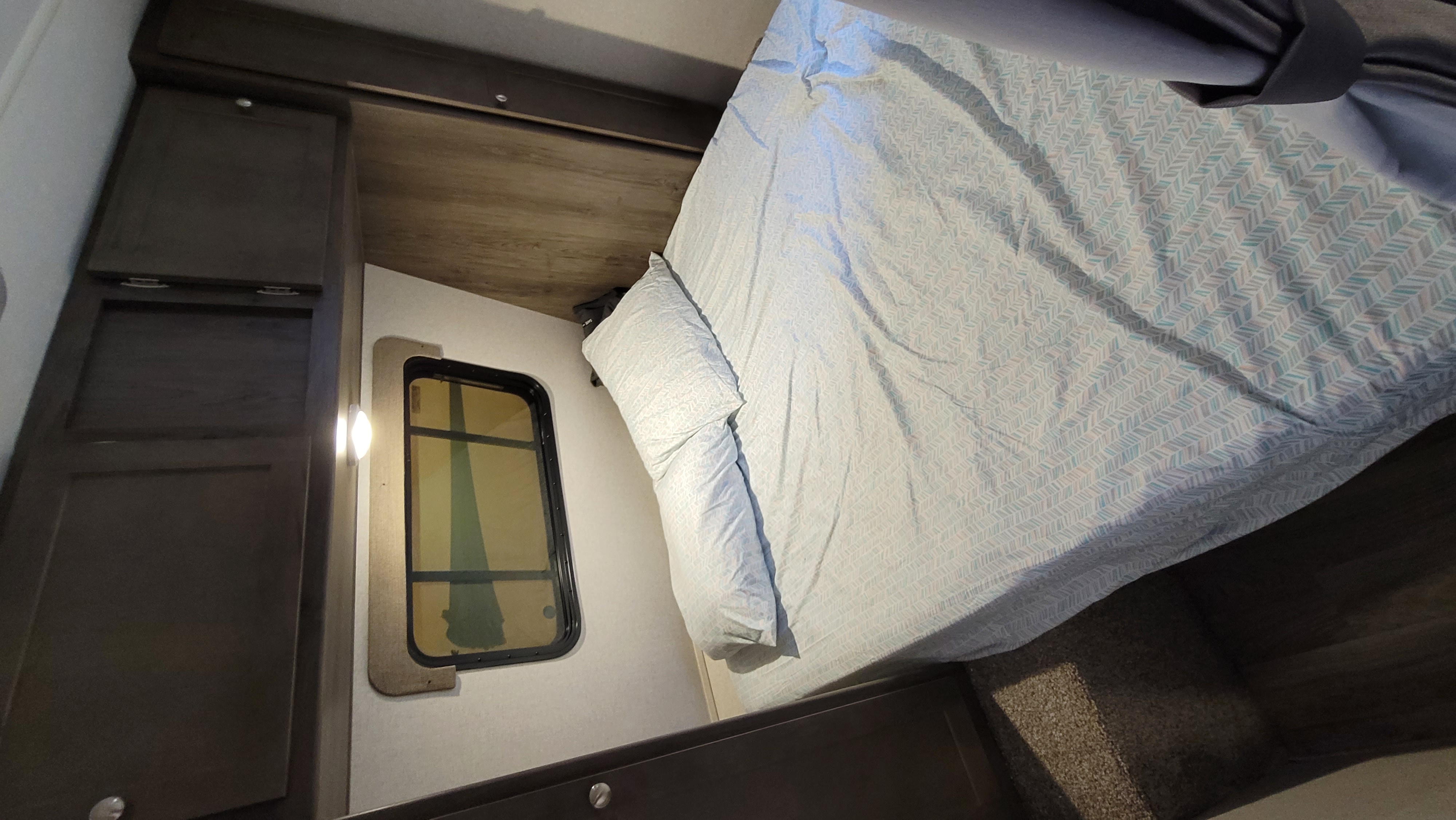 This RV rental bedroom interior features a cozy bed, dark cabinets, and a window with a light fixture for comfortable travel.. Coachmen Viking 2019