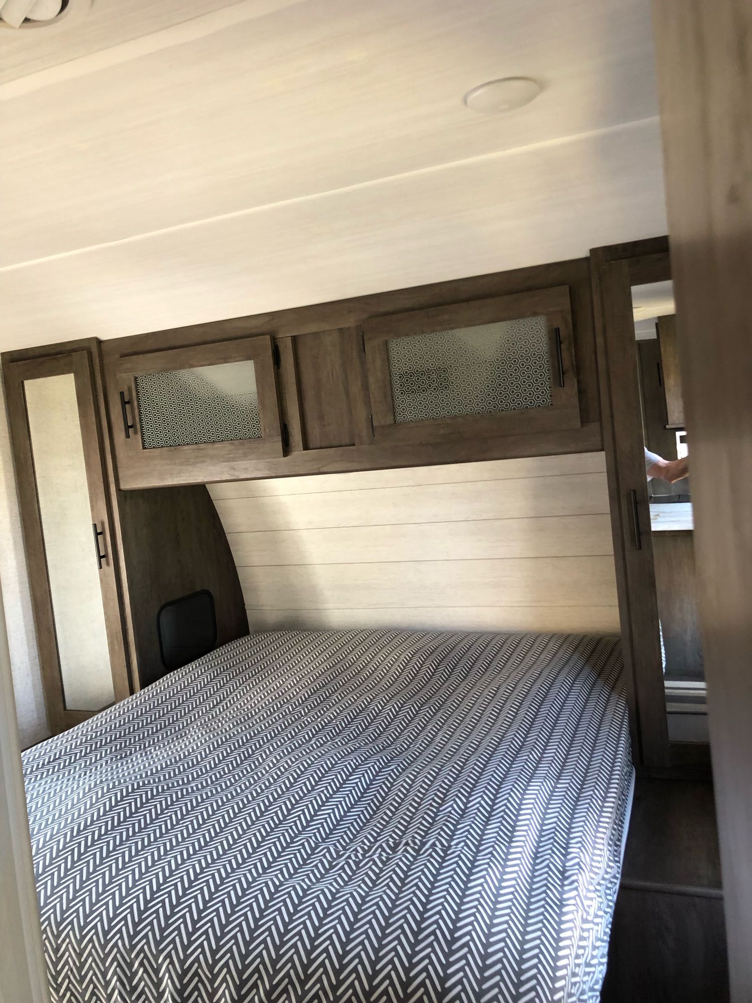 RV rental bedroom: a cozy space with a herringbone-patterned bed, overhead wood cabinets, and light shiplap walls.. Forest River Wildwood 2020