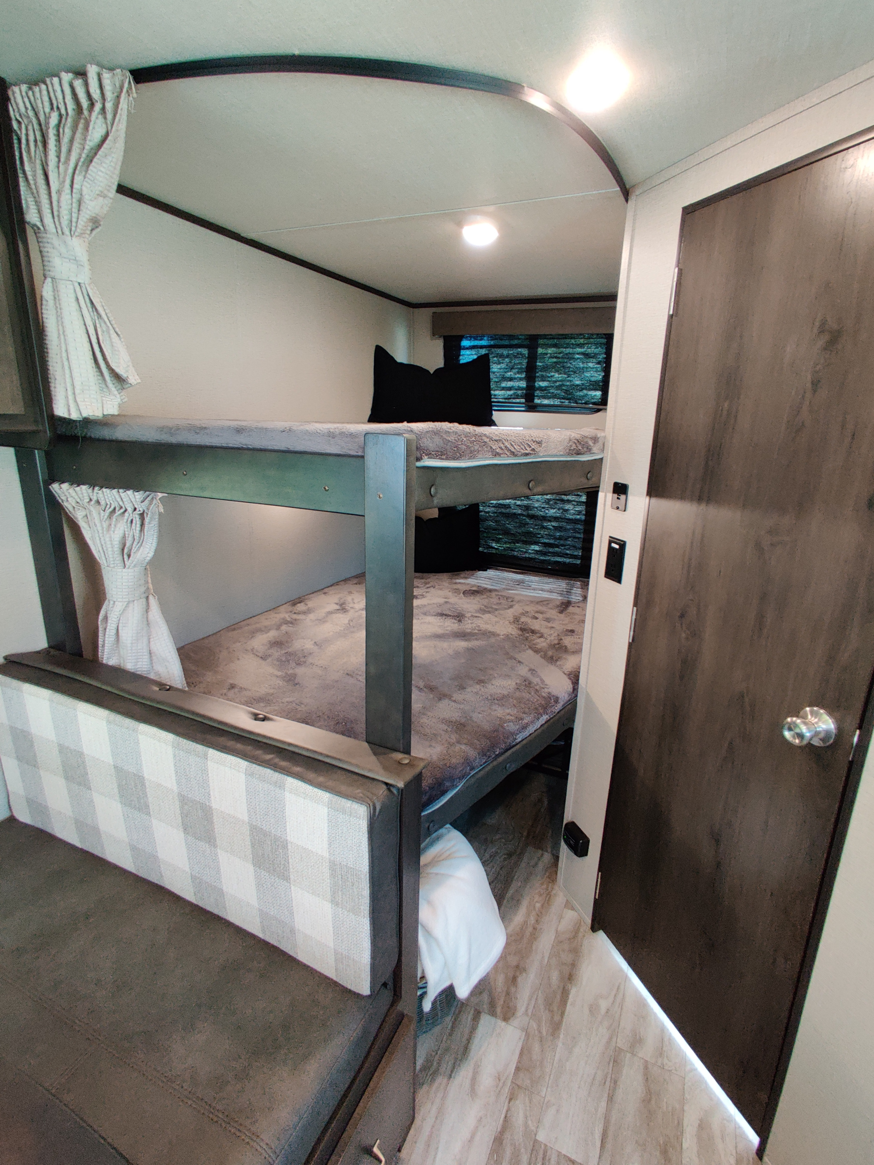 Interior of an rv rental showing space-saving bunk beds with soft bedding, a comfy plaid bench, and modern wood-look flooring.. Grand Design 261BH 2022
