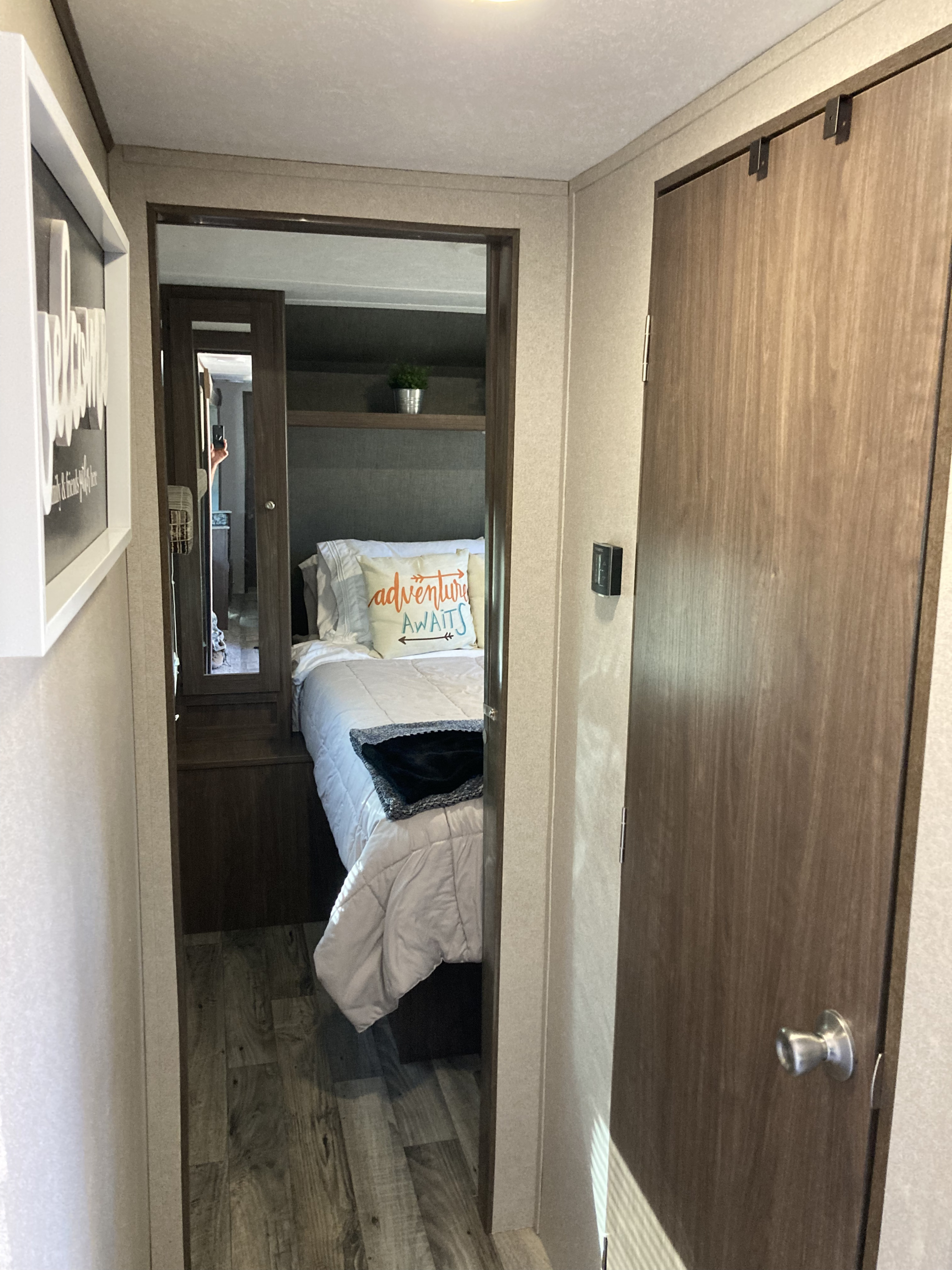 Cozy RV rental interior: A hallway leads to a compact bedroom with a bed, "Adventure Awaits" pillow, and mirror.. Keystone Hideout 2018