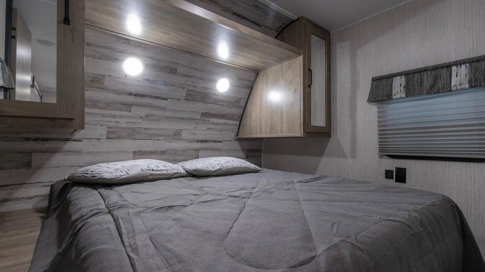 View inside an RV rental bedroom: gray bed, whitewashed wood headboard, overhead cabinets, and soft lighting.. Forest River Palomino Puma XLE Lite 2021