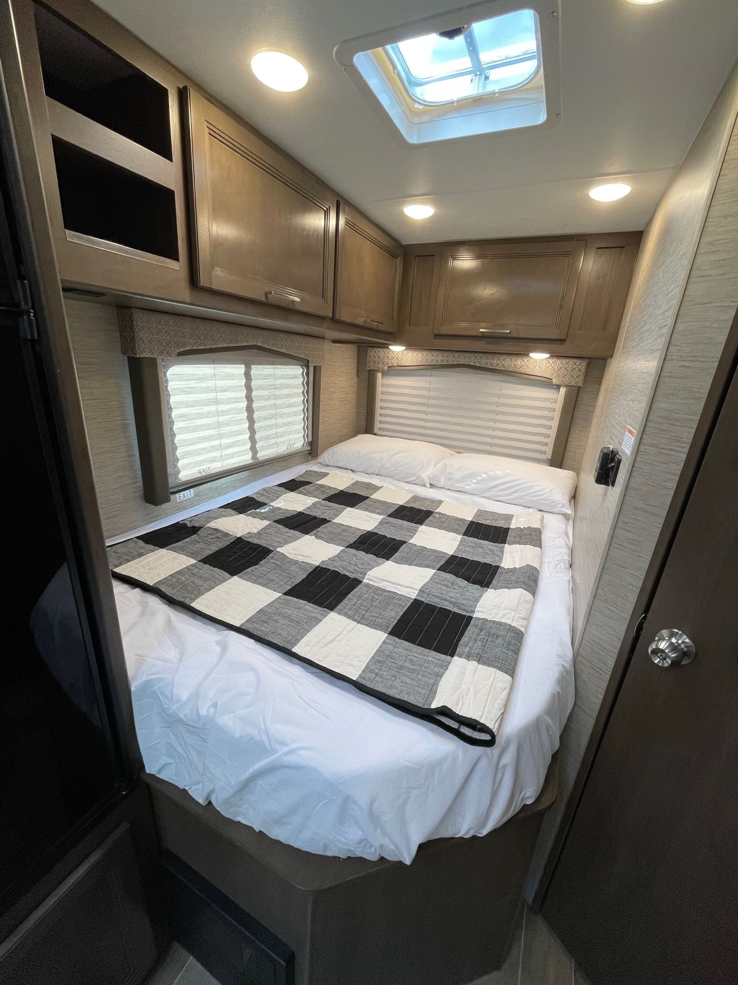 This RV rental bedroom offers a comfortable bed with a checkered blanket, overhead cabinets, windows, and a skylight.. Thor Chateau 2022