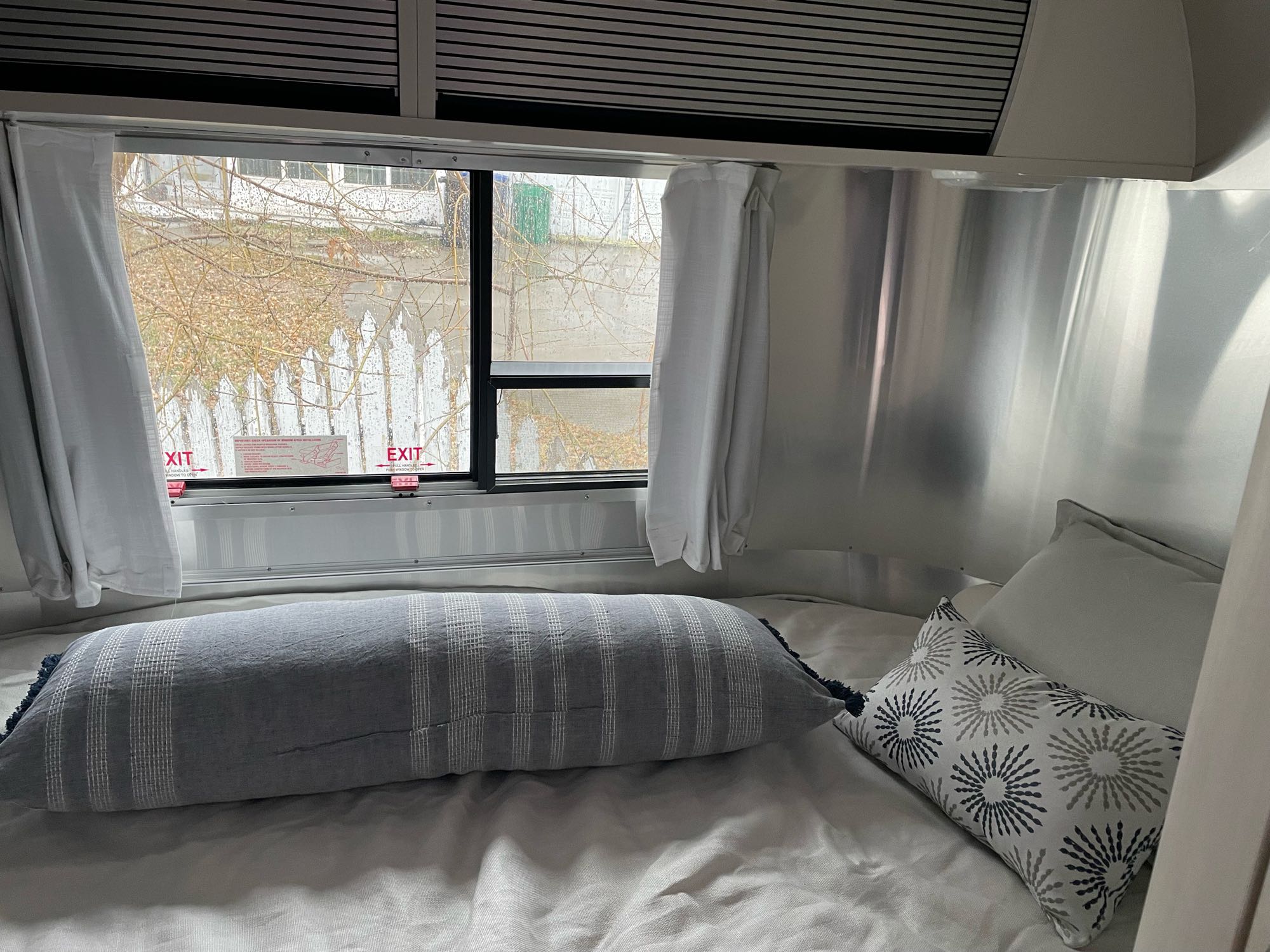 Inside an RV rental: a comfy Airstream bedroom with metallic walls, bed, striped pillow, and window view with emergency exits.. Airstream Bambi 2021
