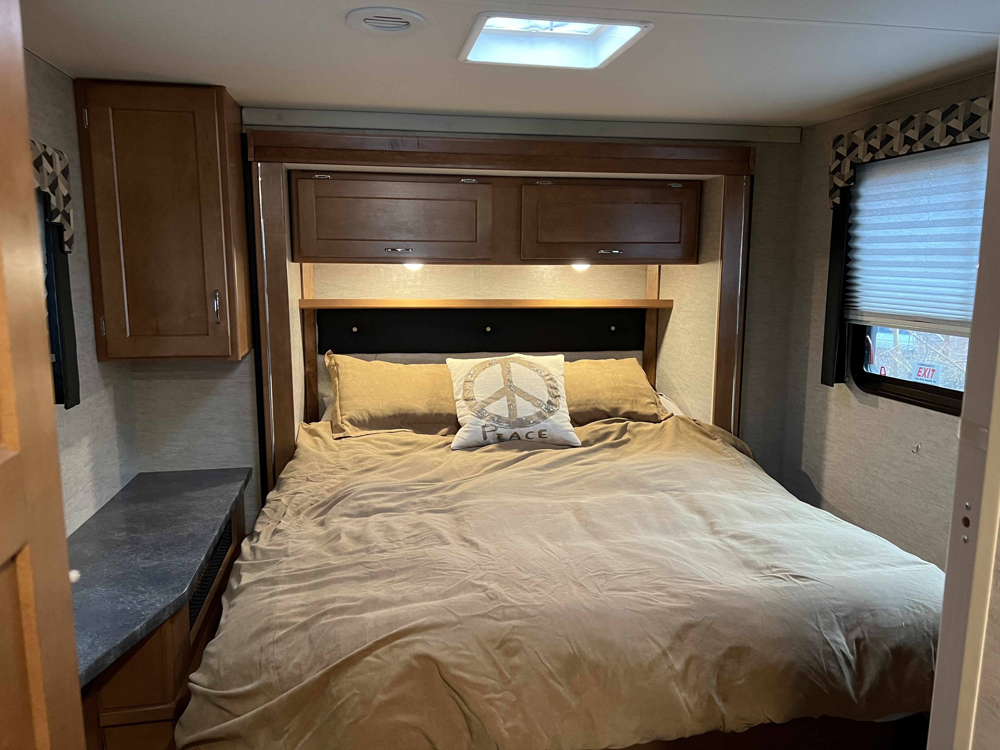 A cozy RV rental bedroom awaits with tan bedding, a peace sign pillow, wooden cabinets, and a window.. Winnebago Minnie Winnie 2017