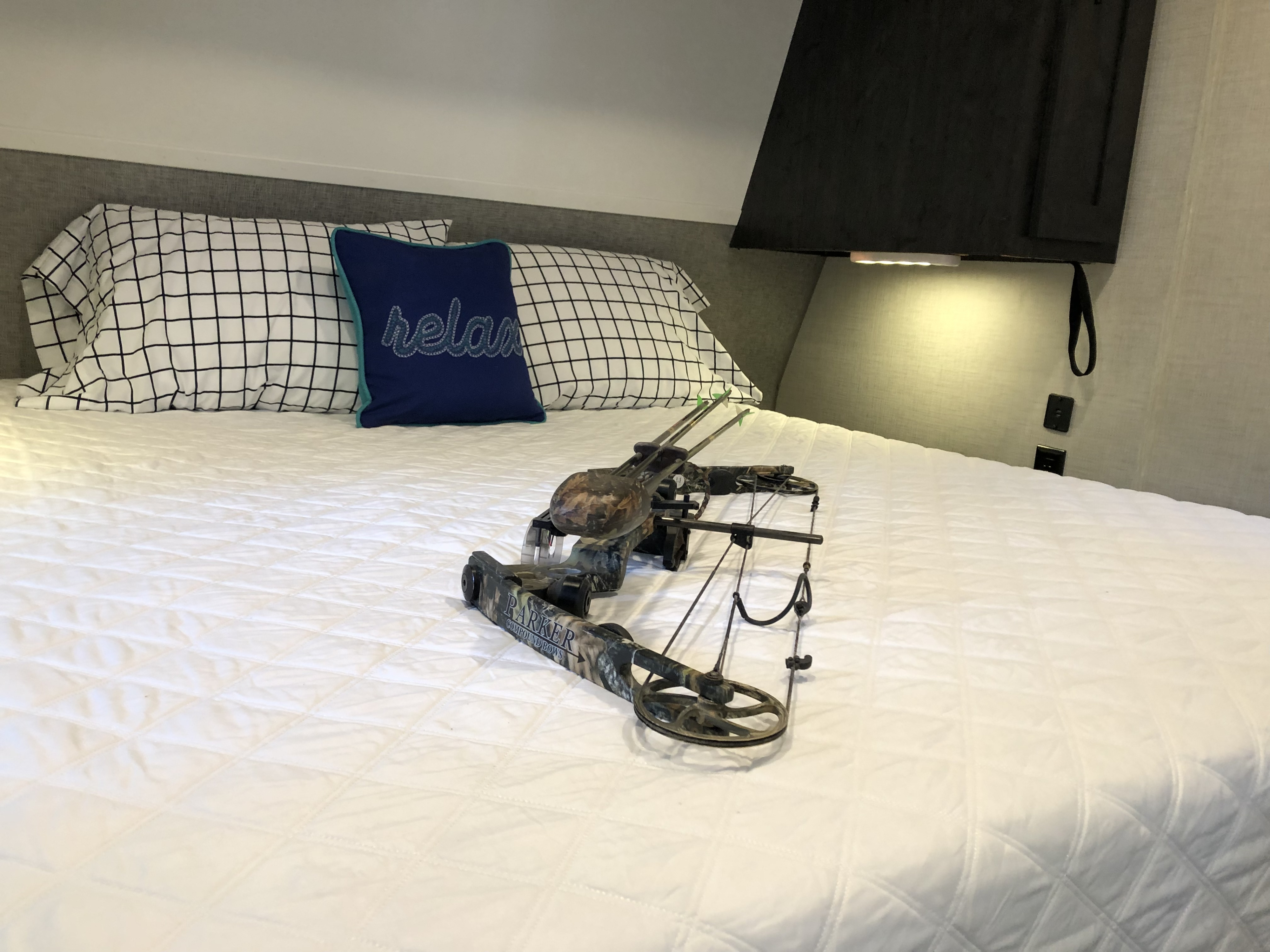 Relax in your cozy RV rental bed! This scene shows a camouflage compound bow on a white quilted bed with grid pillows.. Forest River Ozark 1800 QS 2020