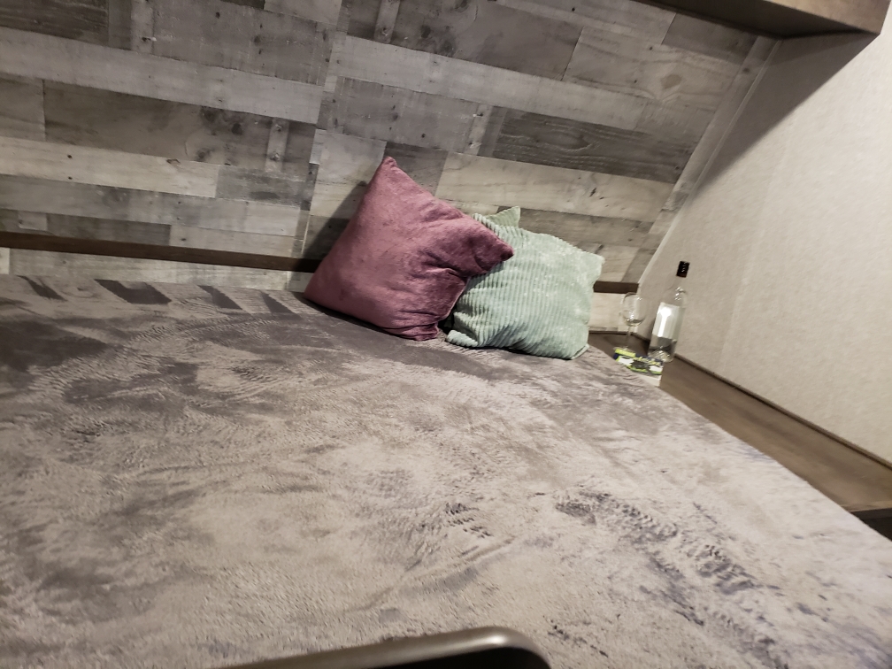 Cozy RV rental bedroom features a bed with plum and green pillows. A water bottle and wine glass rest on a side shelf.. Dutchmen Coleman Lantern LT 2022
