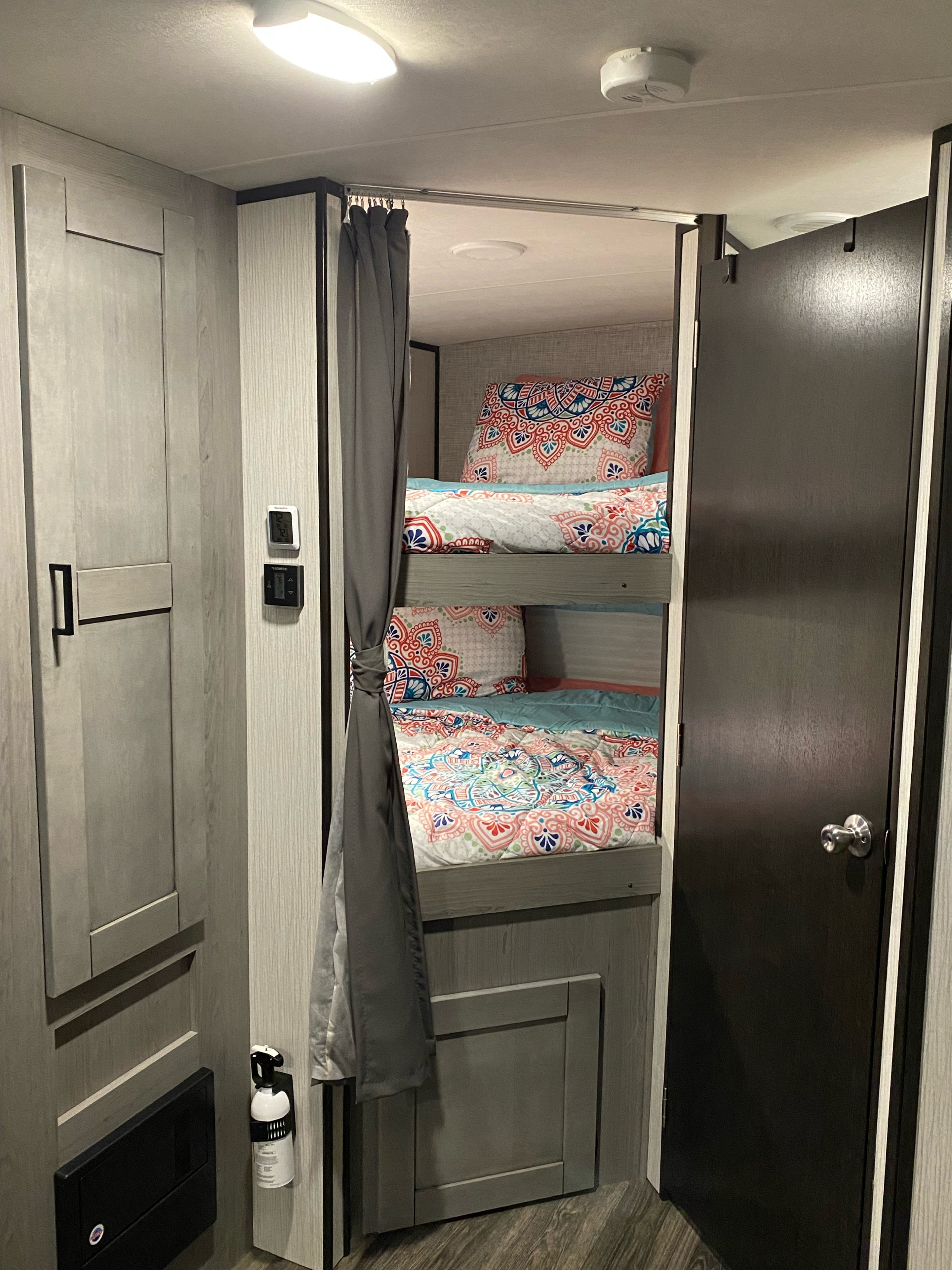 Inside an RV rental: cozy bunk beds with bright bedding, grey privacy curtains, and safety features like a smoke alarm and fire extinguisher.. Heartland RVs Mallard 2021