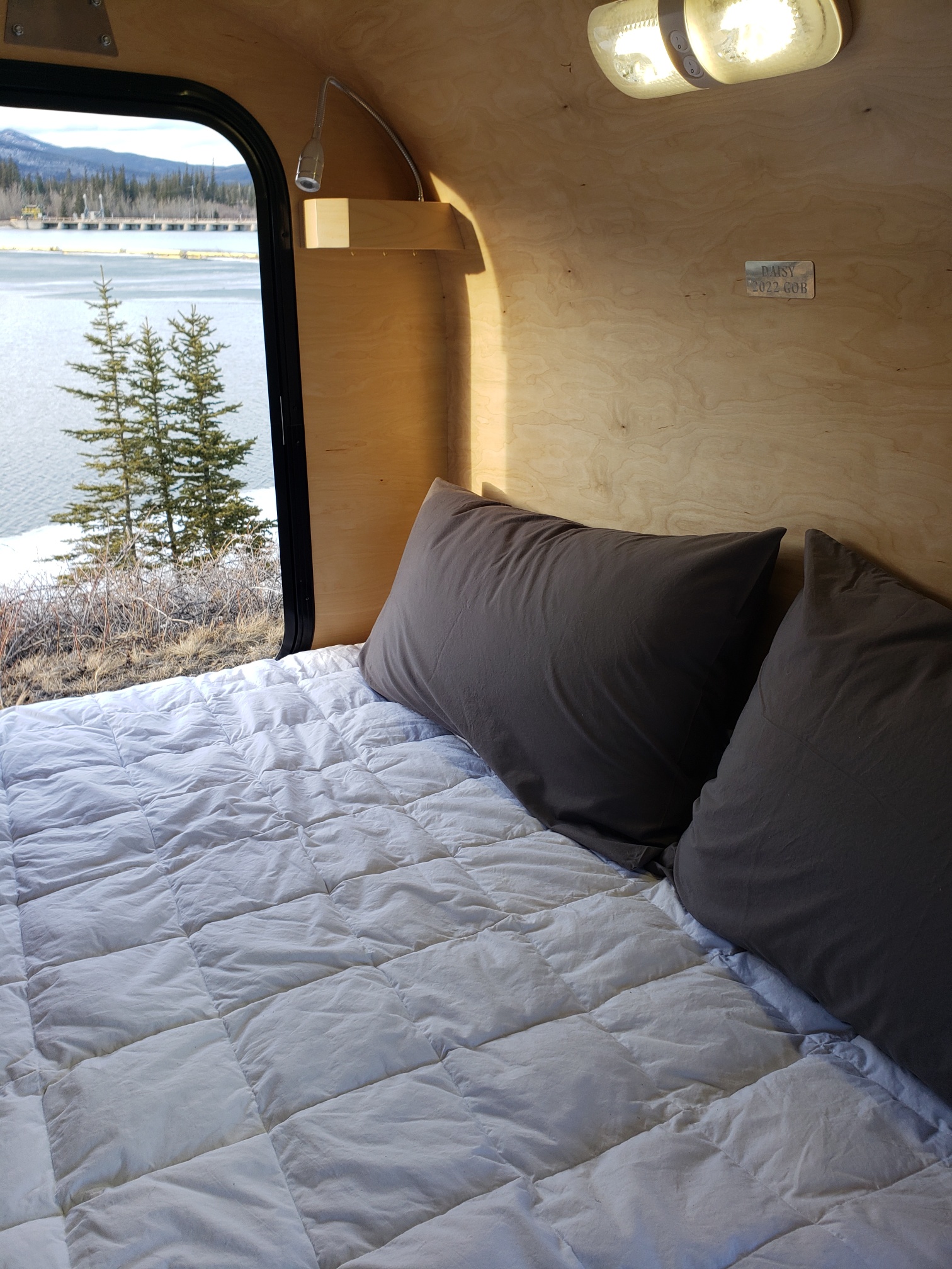 Enjoy this RV rental's cozy bed with a white comforter & a serene lake view through the window, featuring trees & a dam.. Gnome Home Gnome Home 2022