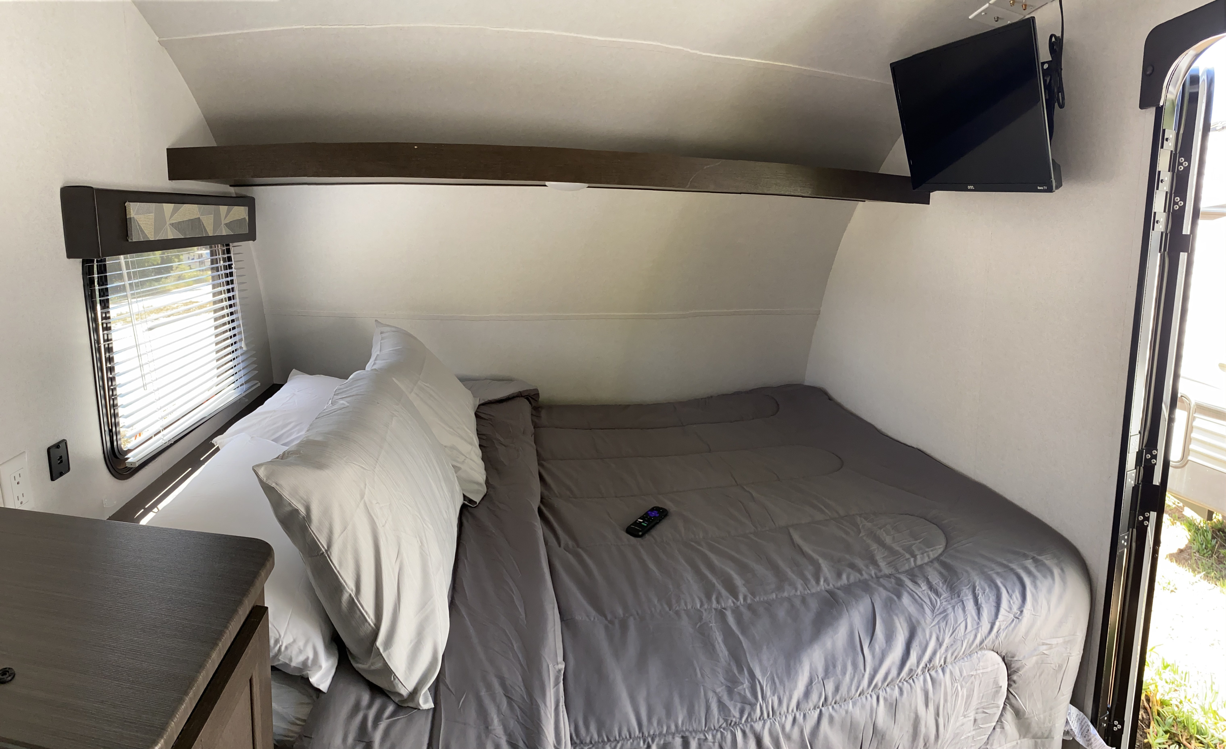 View of a compact RV rental bedroom with a bed, wall-mounted TV, and window with blinds.. Forest River EVO 2020