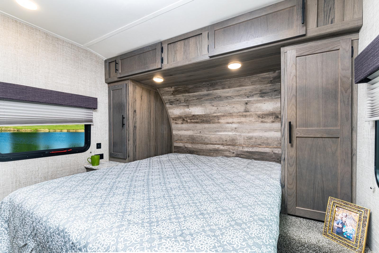 Relax in this cozy RV rental bedroom, featuring a queen bed with patterned bedding, built-in storage, and a scenic lake view window.. Gulfstream 26BHG 2022