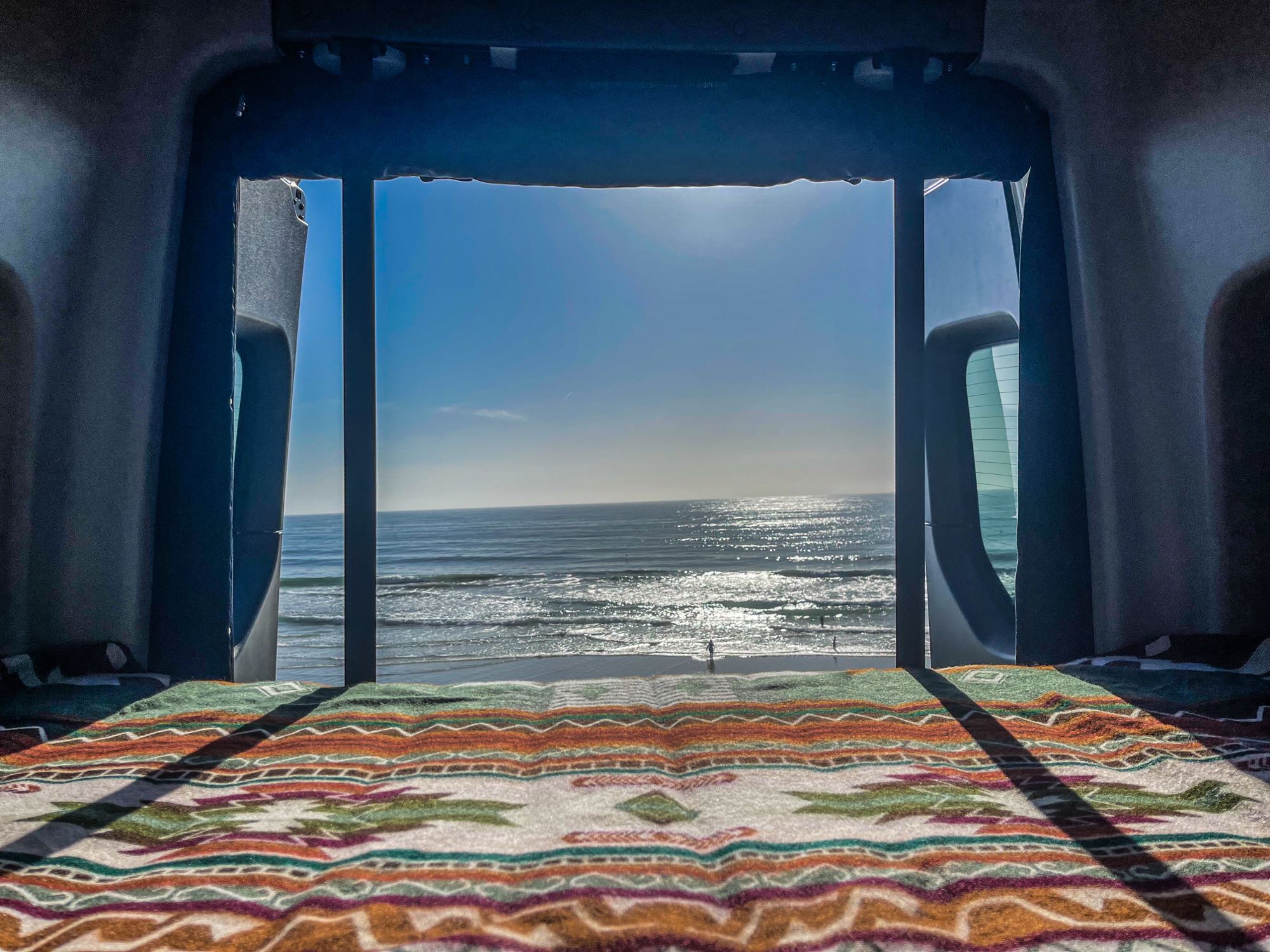 A serene ocean view from inside an RV rental. A patterned blanket covers the bed, offering a cozy spot to watch beach waves.. Winnebago Revel 2022