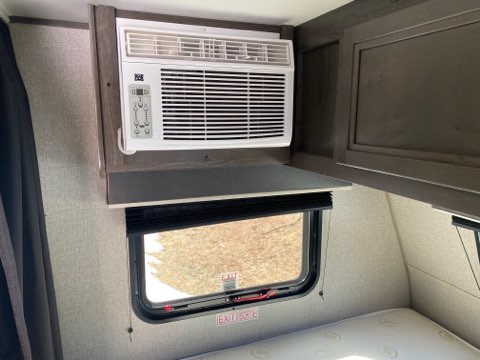 Interior of an RV rental: a compact AC unit mounted above a window, with a small shelf below it, and a bed in the sleeping area.. Jayco Jay Flight 2020