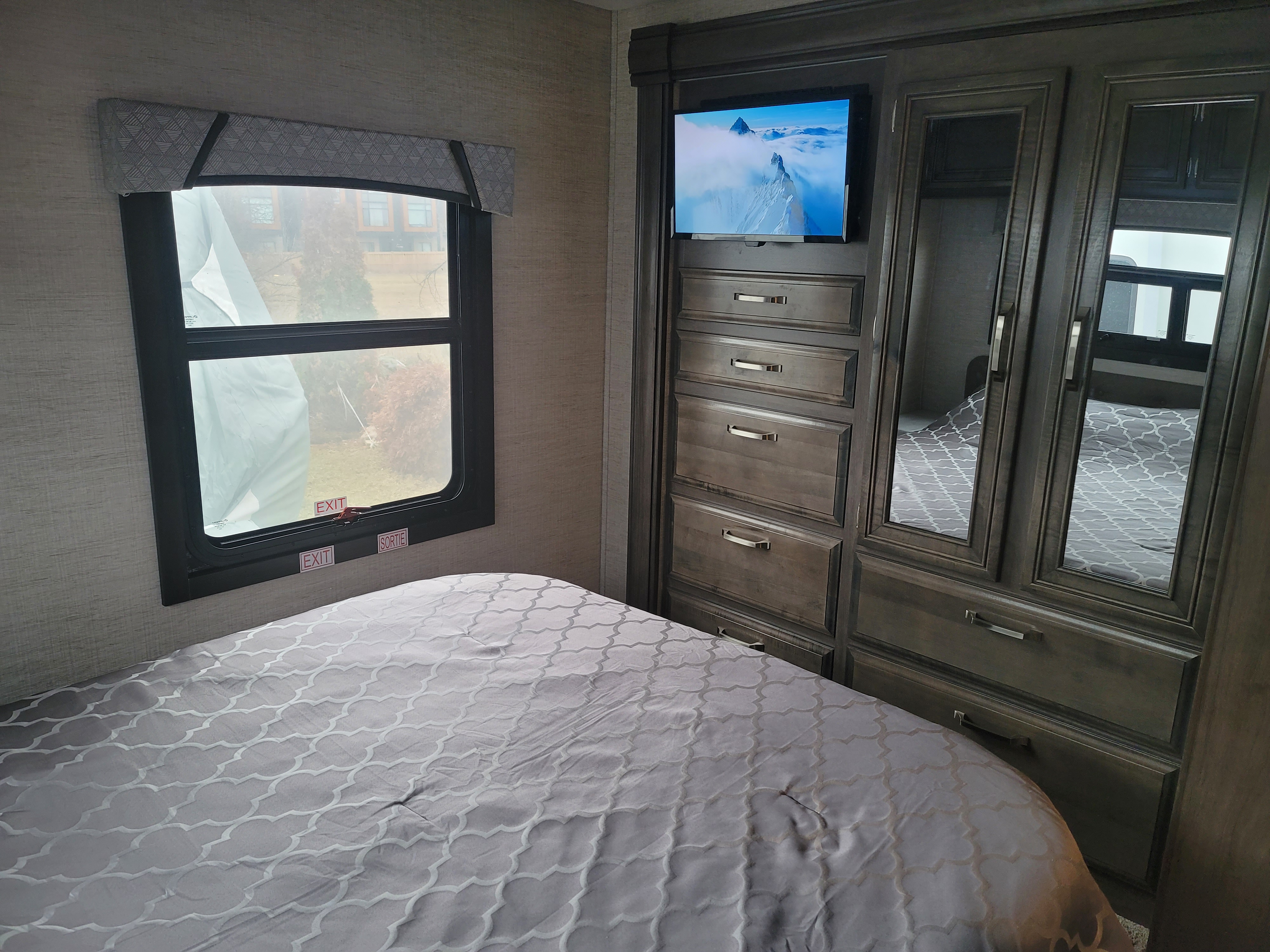 Cozy RV rental bedroom with a patterned bed, emergency exit window, and a built-in wardrobe featuring a TV.. Jayco Prestige 31FP 2022